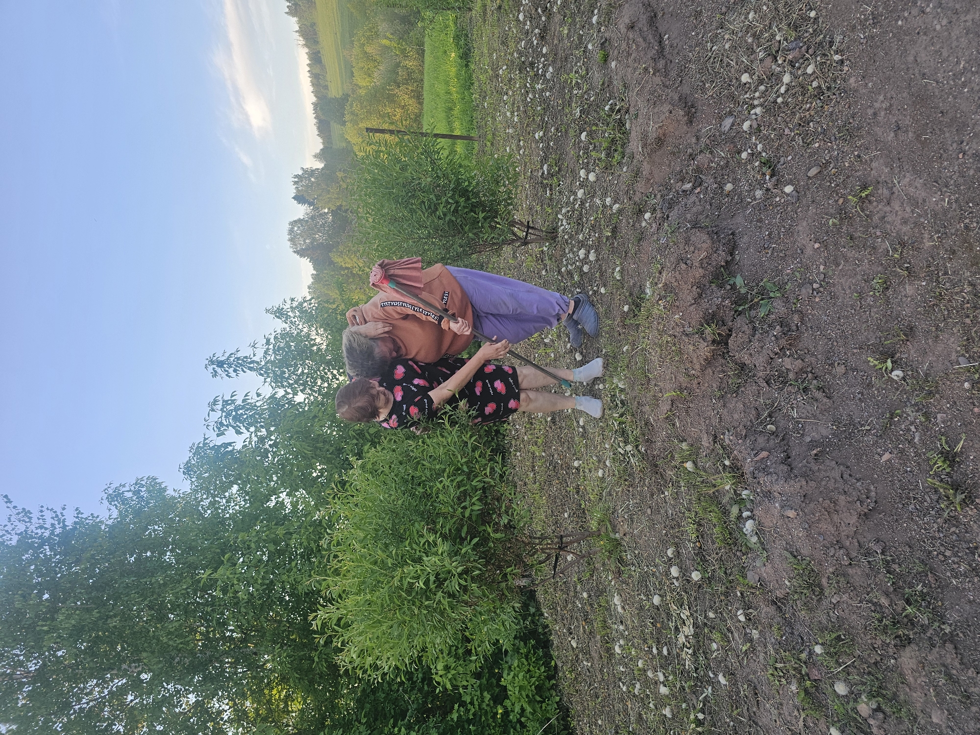 Two elderly women are in an outdoor location near what appears to be a rural area. One woman is wearing a black dress with a red and pink heart pattern, and the other is wearing a brown sweatshirt and purple pants. They are standing near a bush. The ground is a mix of soil, grass, and small stones. The background shows green vegetation and trees, and the sky is visible in the upper left corner. The image suggests a rural setting with natural elements. It's unlikely to pinpoint the precise location within Kalinovka, Russia without further information or landmarks visible in the image.