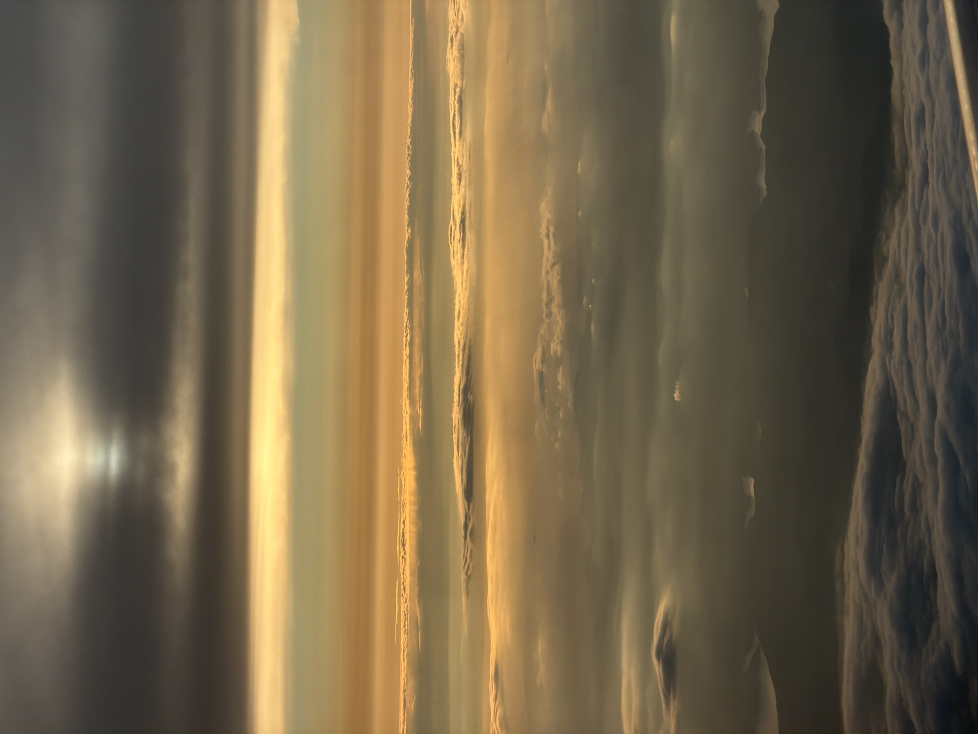 The image presents a view of the sky and clouds, seemingly taken from an elevated position. The sky exhibits a gradient of colors, ranging from dark gray and overcast on the left side to a golden hue towards the center and right. There are distinct vertical bands or layers of clouds, with varying textures and densities. The leftmost area appears to be dominated by dense, dark clouds, possibly indicative of an approaching storm or heavy weather. Moving towards the center, there are layers of golden-toned clouds, suggesting the presence of sunlight filtering through. The rightmost part of the image depicts cloud formations with a more textured and rugged appearance, resembling a mountain range or turbulent weather patterns. The overall impression is one of dynamic atmospheric conditions and a dramatic sky display. There is no visible evidence to suggest the location is Elovoe, Russia.