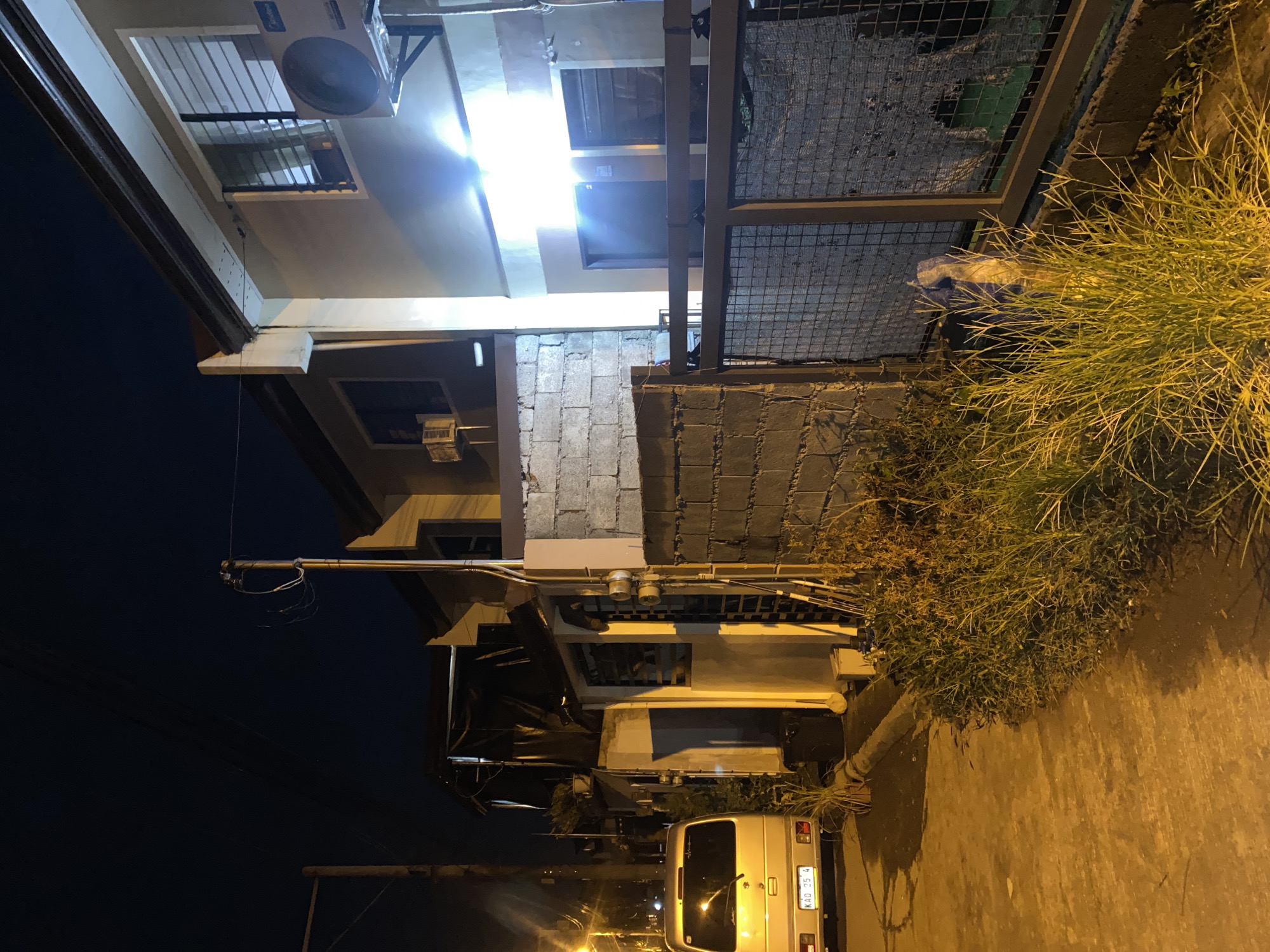 The image depicts a residential street scene at night in Davao City, Philippines. The perspective is from street level, looking along a paved pathway bordered by vegetation and a chain-link fence.
On the left, a silver minivan is parked on the side of the road, facing away from the viewer. Its license plate is visible, reading "KAO 25 4". Adjacent to the minivan is a section of unpaved ground, and further down the street, a utility pole with wires is illuminated by a nearby streetlight.
The right side of the image is dominated by a row of houses. The closest structure is a two-story building with light-colored walls. A prominent air conditioning unit is mounted on the exterior wall. The windows on this level are barred. The house has a dark-colored roof. Another, older-looking house is visible further back, partially covered with a dark tarp. A wall constructed from grey concrete blocks separates the street from the properties. A metal gate with a coarse mesh pattern, partially obscured by overgrown grass, encloses a portion of the property.
The overall lighting is artificial, cast by streetlights and lights from within the houses, creating a warm, yellow-orange hue across the scene. The sky is dark, indicating nighttime. There are no people visible in the image. The environment appears to be a typical urban or suburban residential area, characterized by modest housing, utility infrastructure, and the presence of vehicles.