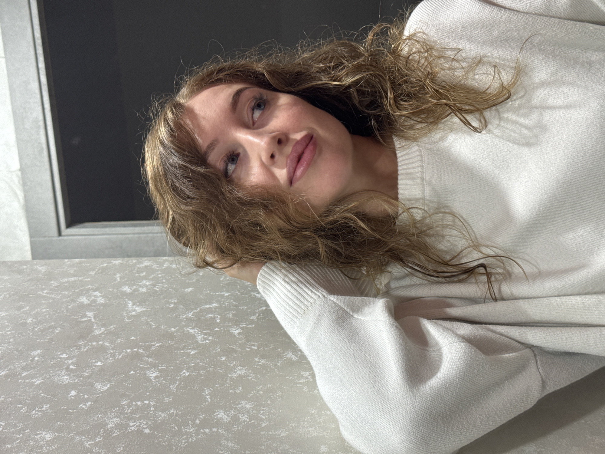 A woman with curly brown hair and a white sweater is resting her head on a gray countertop. The background features a window frame and a dark gray or black wall. The scene is indoors. No further information identifies a specific location within Chaikovsky, Russia.