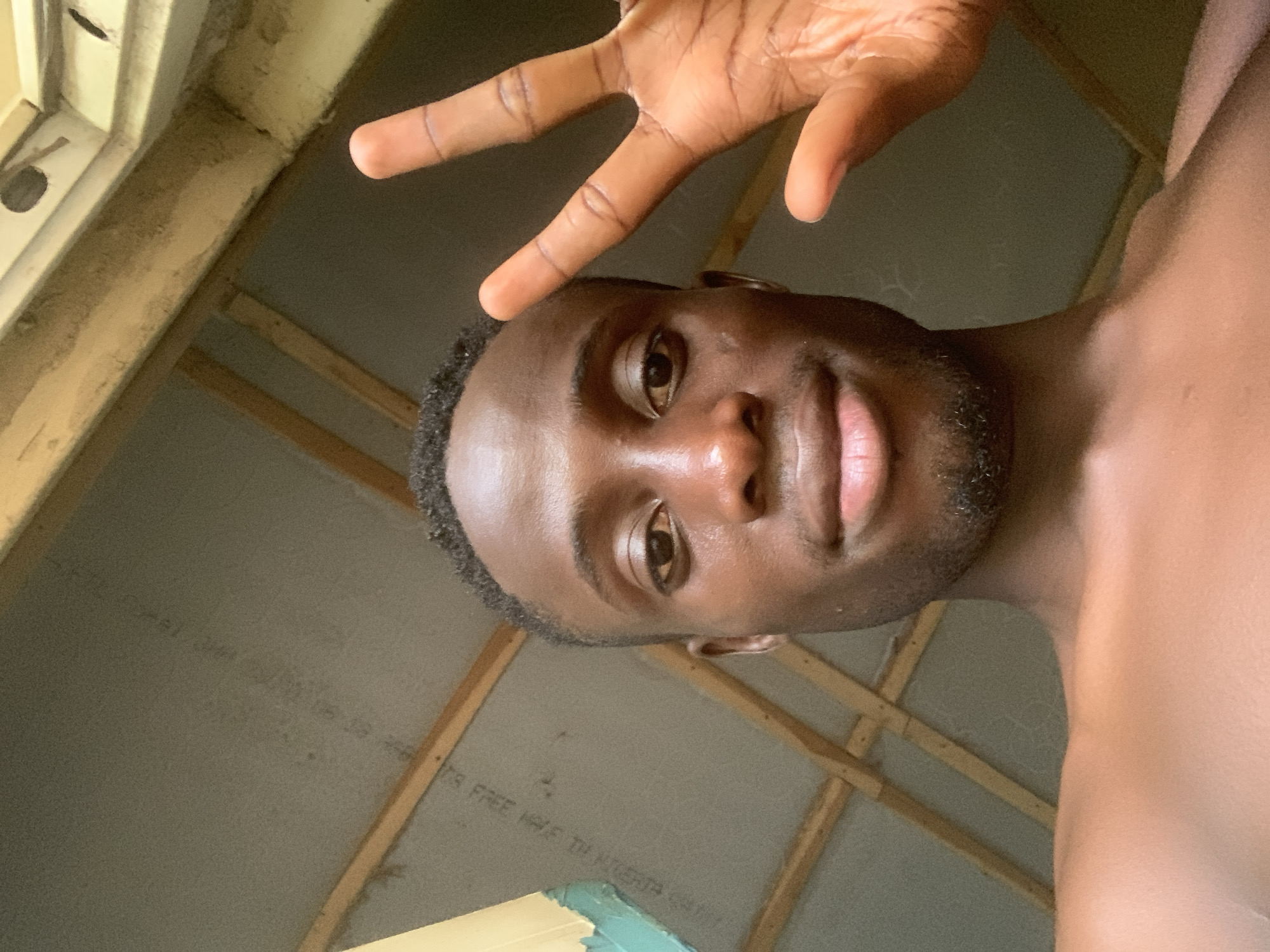 A man is posing for a selfie indoors. He is dark-skinned, with a close-cropped haircut and short facial hair. His right hand is raised above his head in a "peace" sign. The background is a room with exposed wooden beams and grey paneling on the ceiling. There appears to be writing on the paneling. A window is visible in the upper left corner. There is no specific information to confirm this image was taken in Gbeleko, Nigeria.
