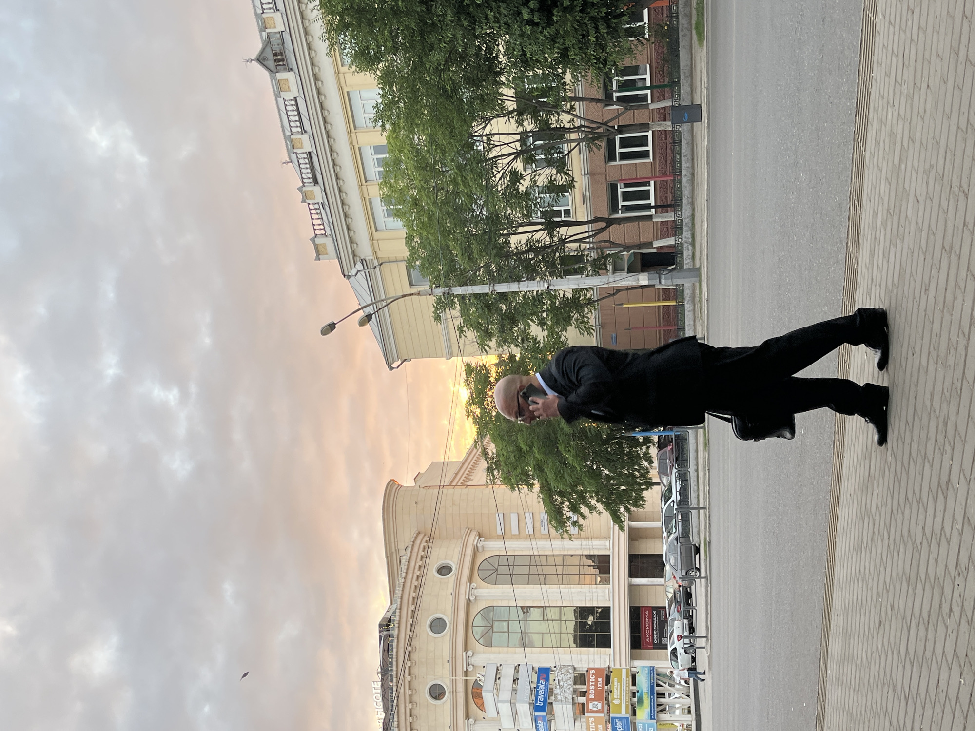 The image shows a bald man in a black suit and glasses walking on a sidewalk and talking on his phone. He is carrying a black briefcase. In the background, there are buildings, trees, streetlights, and cars. The sky is cloudy with hints of sunset colors. The scene appears to be in a city, likely Astrakhan, Russia, given the Russian text on some of the signs in the background, such as "АКСИОМА" (AKSIOMA) and "ОФИС ПРОДАЖ" (SALES OFFICE). The man is the central subject, walking away from the viewer's perspective towards the right side of the frame. The environment suggests an urban setting with a mix of commercial and residential architecture.