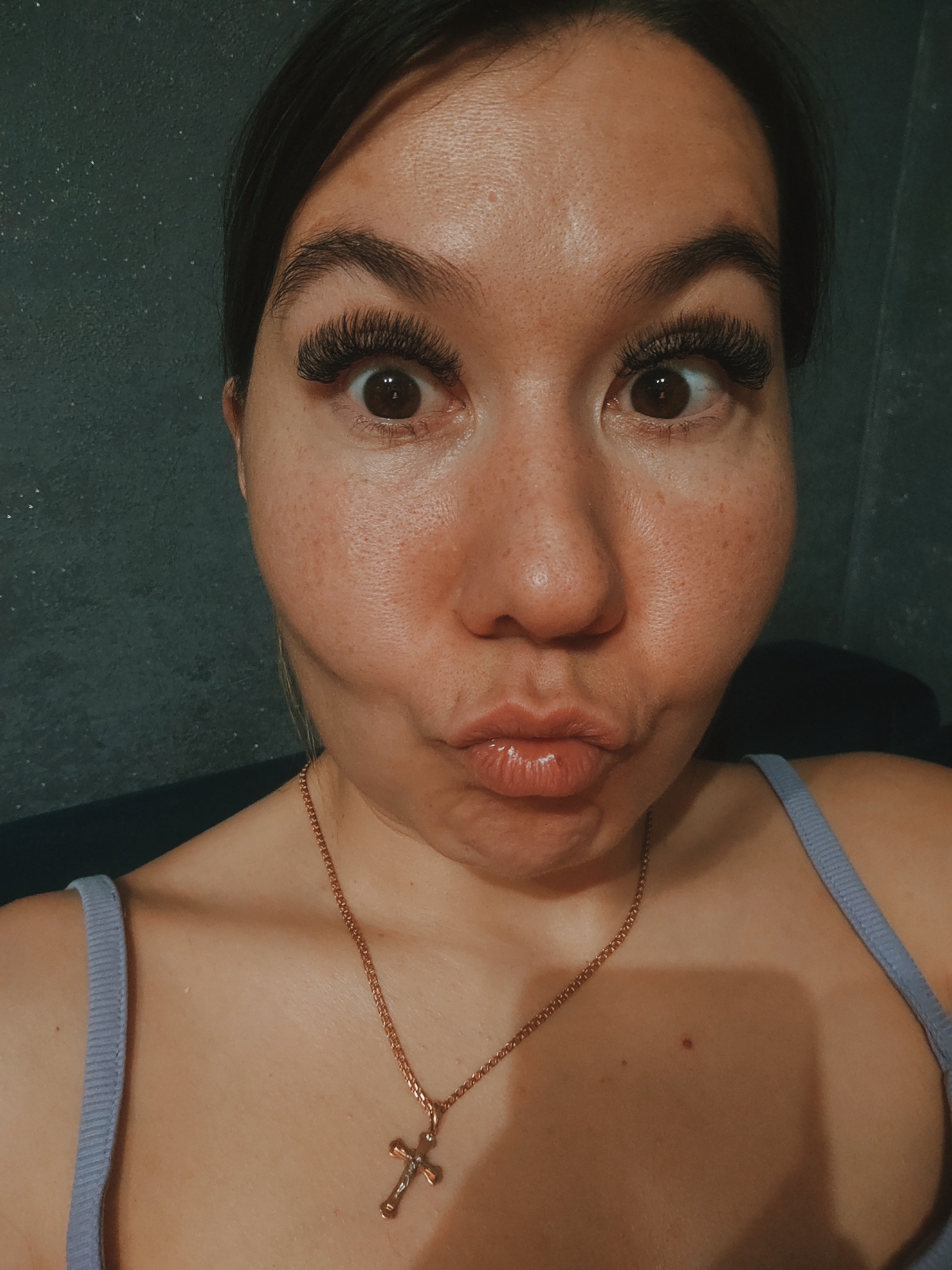 A close-up, front-facing selfie shows a woman with wide-open eyes and her lips pursed in a pout. She has dark hair pulled back, dramatic false eyelashes, and defined eyebrows. Her skin has visible pores and some freckles. She is wearing a thin gold chain with a cross pendant and a light purple tank top. The background appears to be a dark, textured wall. The image is lit from the front, creating a slight shadow on her left cheek.
