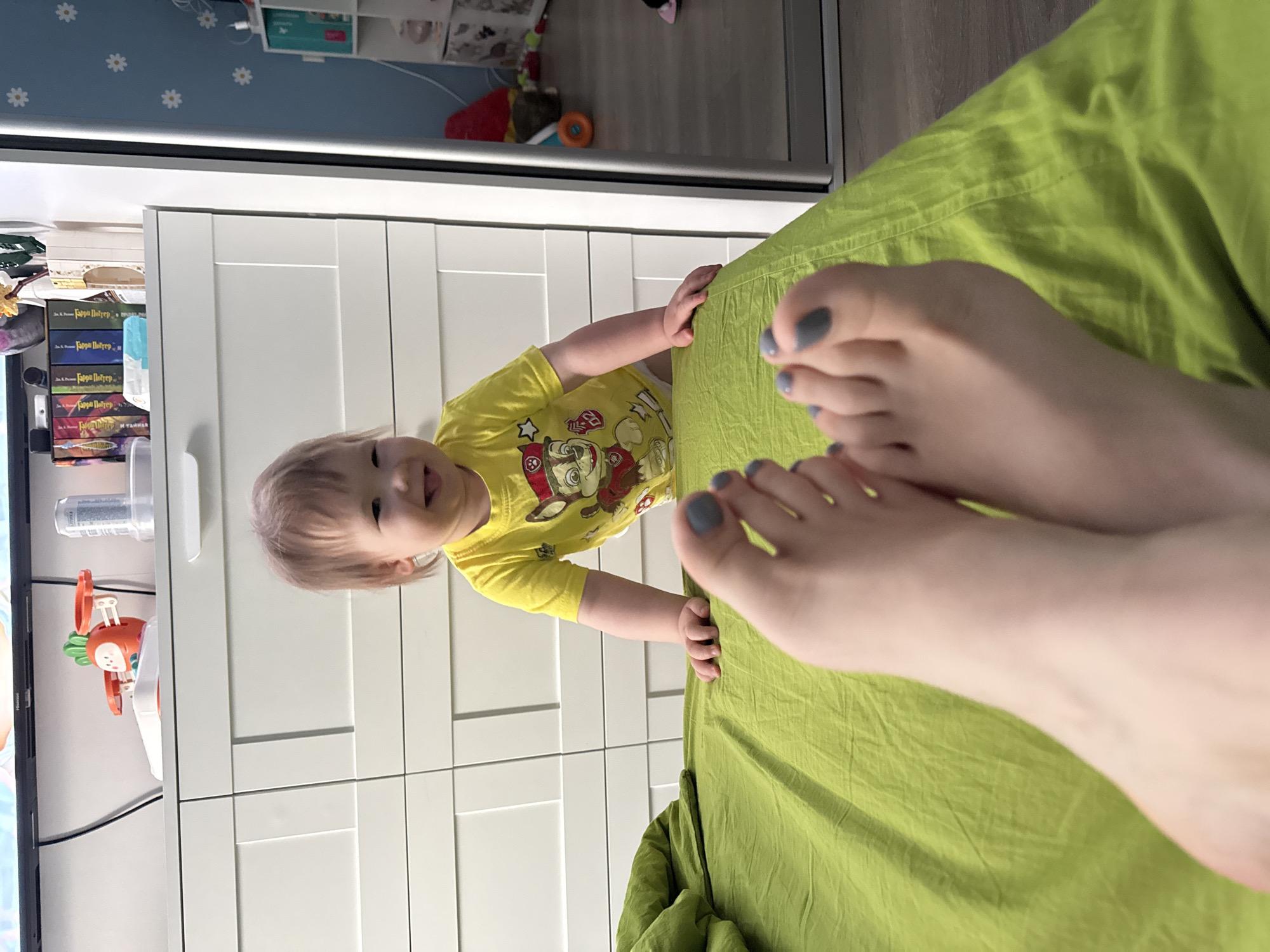 A child in a yellow shirt, visible from the waist up, leans towards a bed covered in green sheets with two feet in the foreground. The feet have grey nail polish. Behind the child is a white wardrobe. To the left is a partially visible TV screen and a bookshelf containing children's items and books, including books with titles in Russian. The location is likely indoors. There is not enough information to determine the location in Moscow.