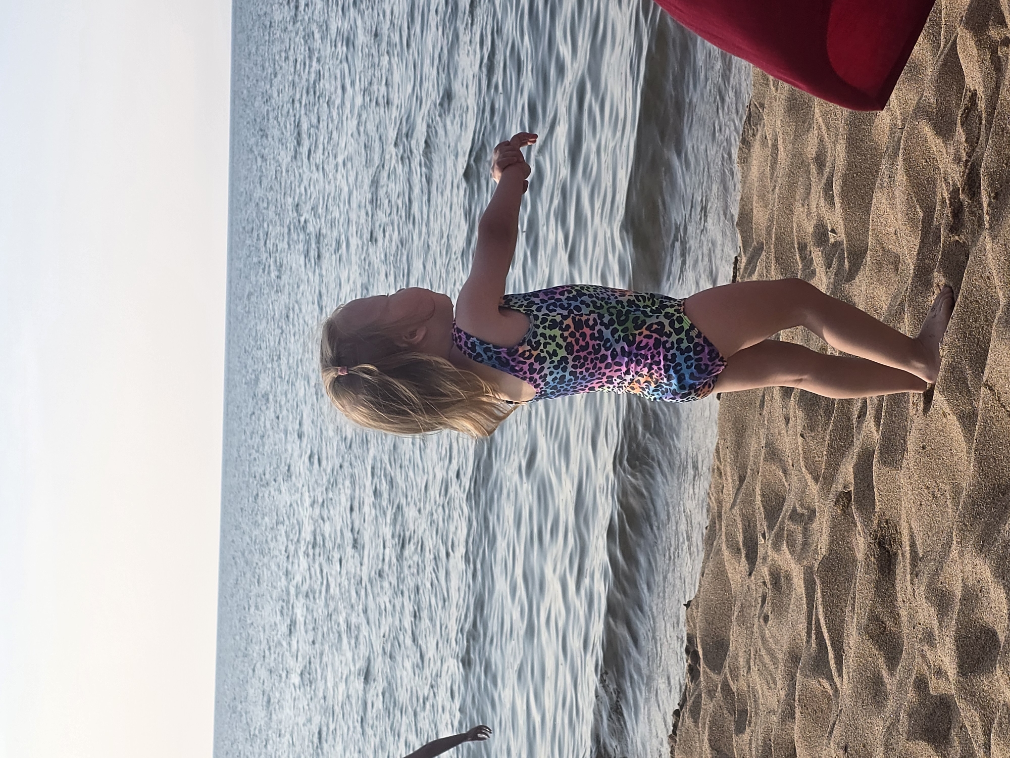In Maksimikha, Russia, on a sandy beach with a horizon of blue water under a clear sky, a young girl in a colorful leopard-print swimsuit stands facing right, her arms outstretched. The girl has blonde hair tied into a ponytail with a pink elastic. In the background, the calm, rippling surface of the water stretches to the horizon. To the left of the girl, a partial view of another person's arm is visible, reaching out towards the water. The sand in the foreground is textured with ripples and undulations. A sliver of a red object, possibly a towel or umbrella, is visible on the far right edge of the image.