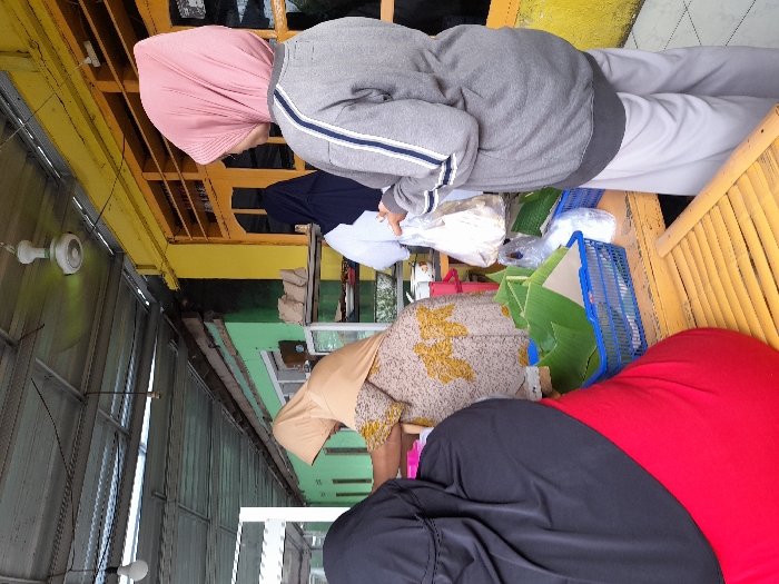 A trio of individuals engages in an activity, likely a transaction, at a roadside establishment. Two are seated, with the one nearest in a red and black outfit, the other adorned in a patterned tan garment. A third figure stands, cloaked in a gray jacket and pink head covering. A vibrant yellow structure forms the backdrop, with a nearby collection of banana leaves and blue baskets suggesting a food-related business.