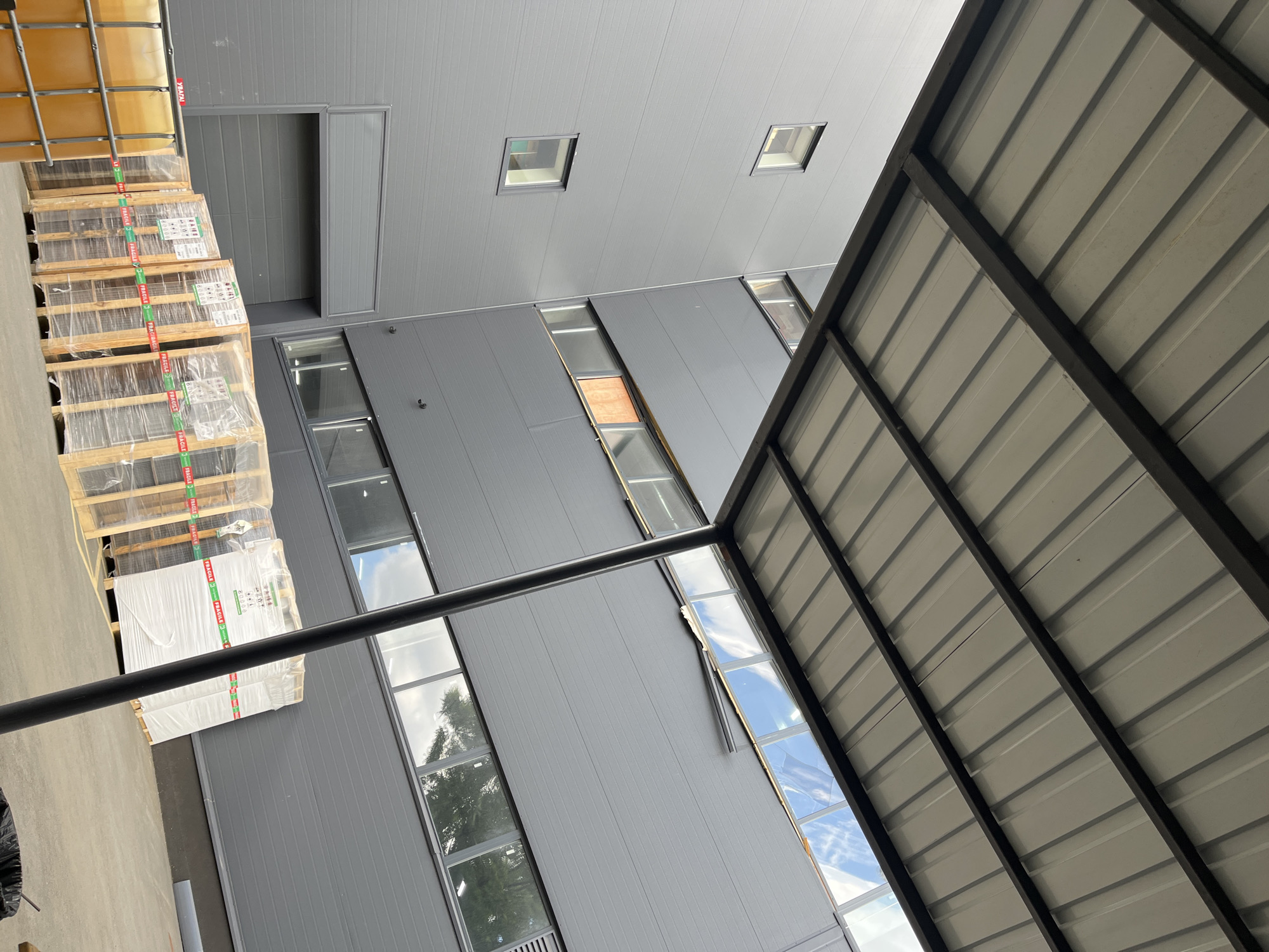 The image shows an exterior view of a modern industrial building with a gray facade, possibly a warehouse or factory, located in Kyiv, Ukraine. A portion of a metal-roofed canopy extends from the building's upper section, supported by a dark, vertical metal pole. The building features a series of horizontally oriented windows, some of which reflect the sky and clouds. In the foreground, there are several pallets of goods wrapped in plastic, with red "FRAGILE" tape. To the left, a large yellow translucent tank, possibly containing liquid, is partially visible. The ground appears to be concrete or asphalt. The scene is devoid of people.