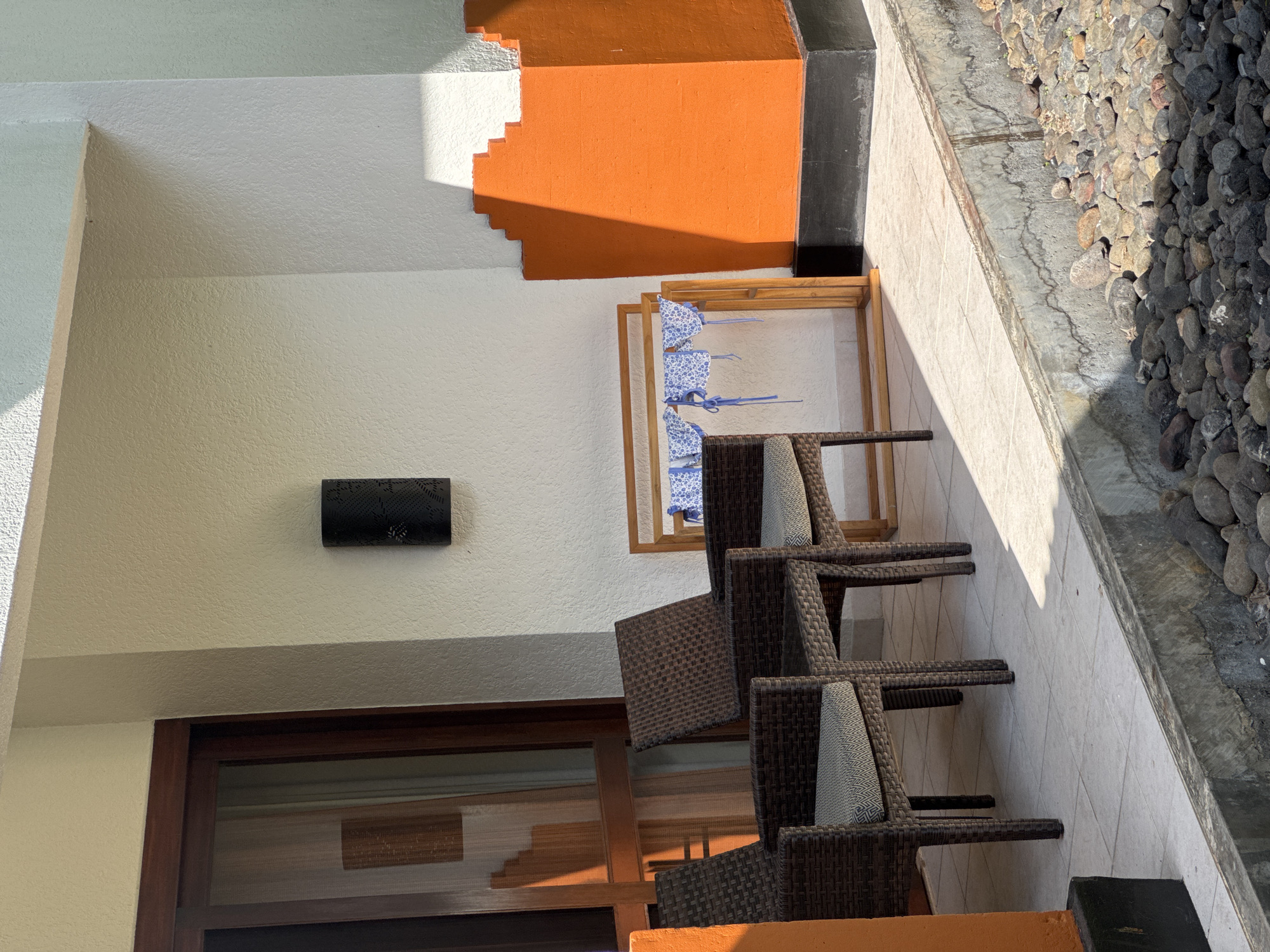 The scene shows an outdoor patio area in Kuta Selatan, Indonesia. Three brown wicker chairs with patterned cushions are arranged in a line on a white tiled floor. Next to the chairs is a wooden rack holding baby bonnets. The patio is adjacent to a wall, painted white, with a section near the top painted orange with a stepped pattern. A small, dark-colored object is mounted on the wall. A window with a dark brown frame is visible on the left. A narrow garden with grey stones borders the patio on the right.