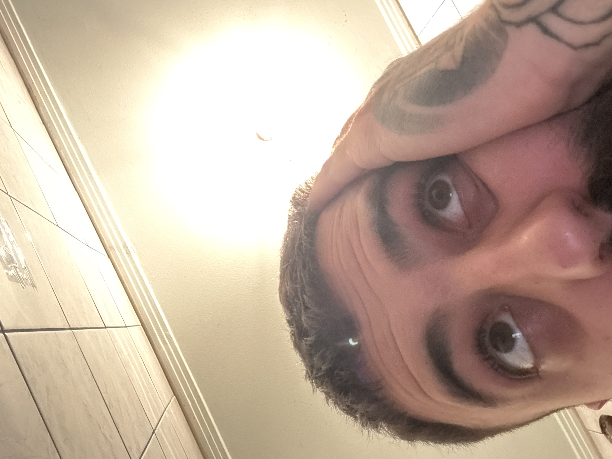 The image displays a close-up, upward-angled perspective of a male individual, located in Vrachesh, Bulgaria. The subject's upper face, including the forehead, eyes, nose, and part of the cheek, is visible. The person has short, dark hair, dark eyebrows, and a visible beard and mustache. Both eyes are open and directed upwards, reflecting a bright overhead light source. The individual's left hand (viewer's right) is positioned against the side of their face, partially obscuring the right temple and cheek. This hand displays extensive dark-ink tattoos across the back of the hand and forearm. The background consists of a light-colored, tiled ceiling in the upper left quadrant, with a textured, light-colored wall below it, separated by a thin horizontal trim. A dominant, intensely bright, circular light source is positioned in the upper right portion of the image, causing significant illumination and lens flares, including a distinct crescent shape near the center of the light source and another smaller one on the subject's forehead.