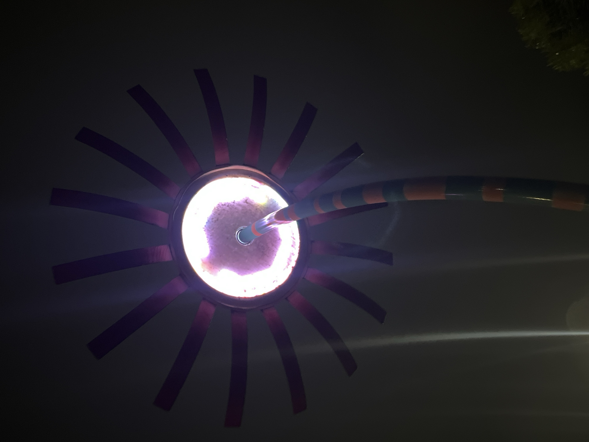 The image displays an illuminated art installation or light fixture at night in Chaikovsky, Russia. The central object is a stylized sun or flower-like light source featuring a circular core emitting a bright, textured pinkish-purple glow. Radiating from this core are approximately 16 dark reddish-purple, elongated, rectangular elements, resembling petals or sun rays. A pipe or pole, horizontally striped with alternating bands of orange and dark green, extends from the lower center of the frame, diagonally upwards, into the glowing core of the fixture. The background is predominantly dark, indicating nighttime conditions. On the lower left, faint vertical light streaks are visible, possibly lens flares or atmospheric reflections. In the bottom right corner, a small section of dark green foliage is discernible. No people are visible in the scene.