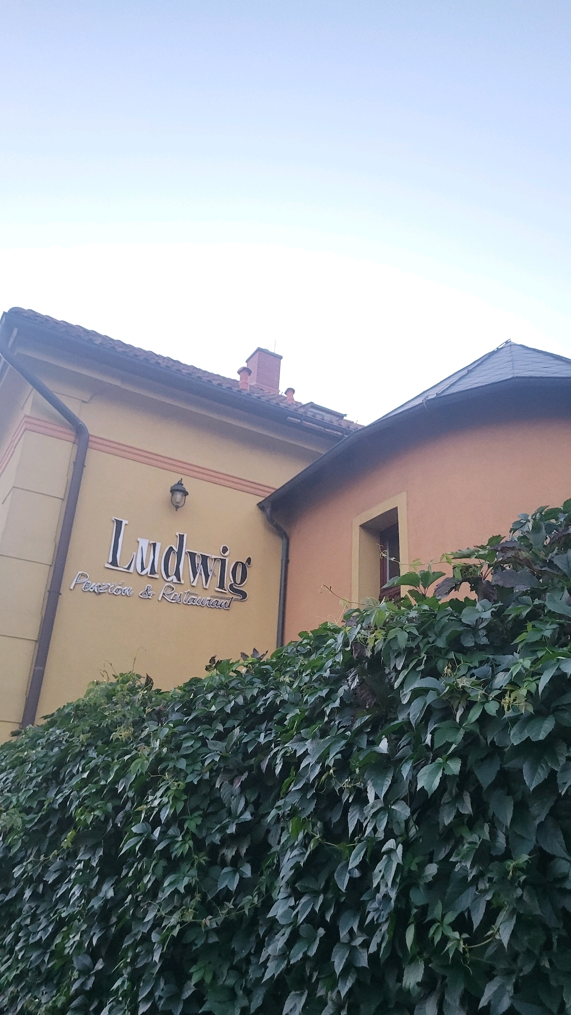The image shows a building with a sign that reads "Ludwig Penzión & Restaurant". The building is colored yellow and orange, and is covered in ivy. The sky is visible in the background. The building is located on Floriánska street in Prešov, Slovakia.
