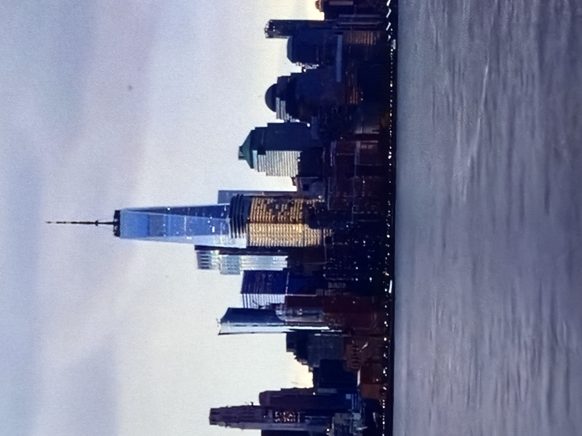 The image displays a panoramic view of a major cityscape dominated by numerous skyscrapers, seen across a body of water. The most prominent structure is the One World Trade Center, distinctly identifiable by its height, reflective blue glass facade, and antenna spire, positioned centrally within the skyline and illuminated with a blue hue towards its upper sections, transitioning to warmer tones lower down. Surrounding it are various other high-rise buildings, some displaying illuminated windows with yellowish light, while others appear as darker silhouettes against the sky. The city's waterfront shows a line of numerous small, bright lights, likely streetlights or building lights. The foreground features a wide expanse of calm, dark blue-grey water, showing subtle ripples and hints of reflection. The sky above is overcast, presenting a soft, muted grey-white tone, suggesting either dawn or dusk. No people are visible.
This image depicts the skyline of New York City, United States, specifically Lower Manhattan, as viewed from across the Hudson River or New York Harbor. The location specified in the prompt, Maidstone, United Kingdom, is incorrect; the image does not show Maidstone.
