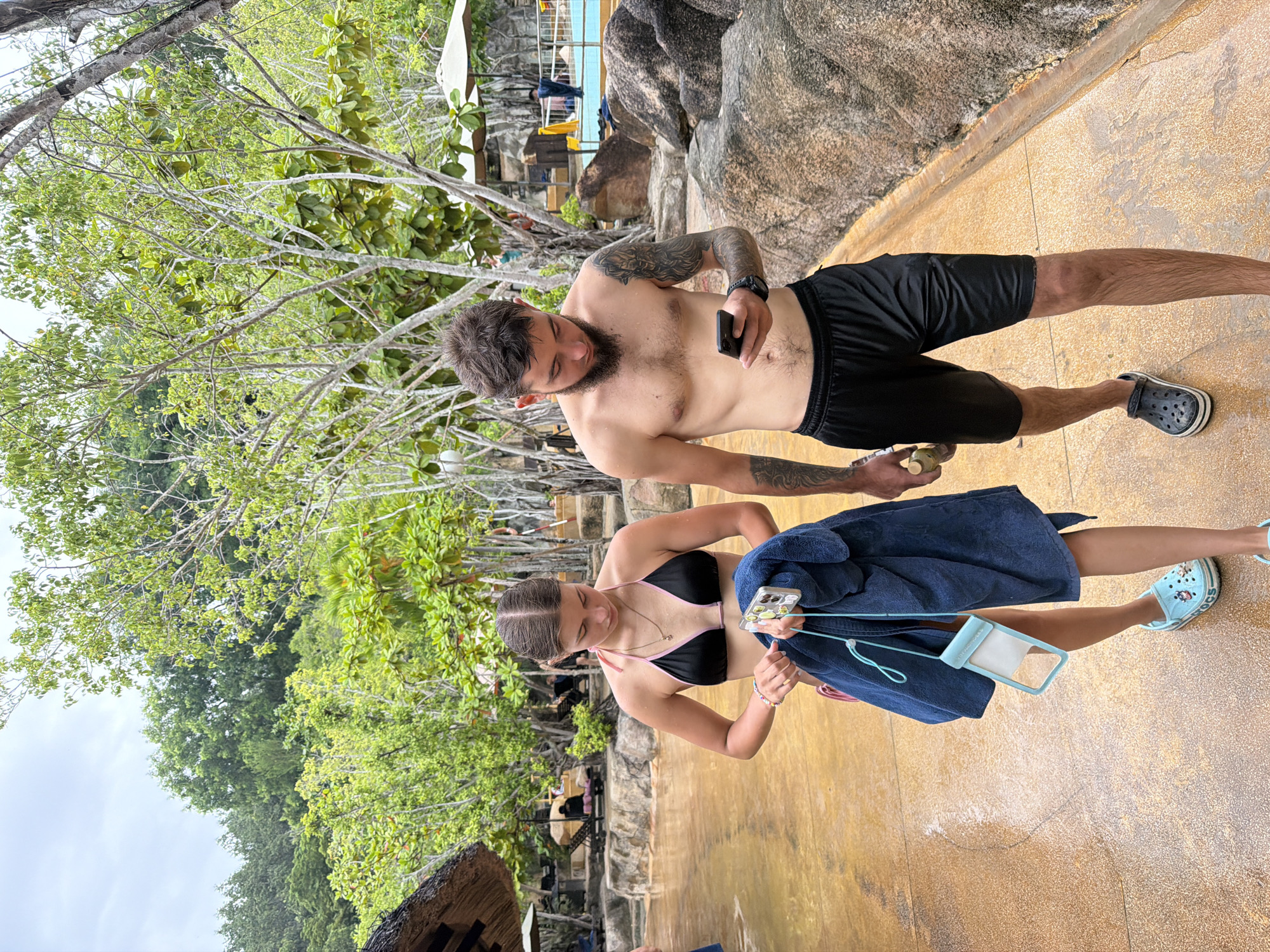 In Nha Trang, Vietnam, a young couple walks along a wet, sandy pathway next to a swimming pool area. The female, on the left, wears a black bikini with pink trim, holding a dark blue towel and a clear waterproof phone pouch with a blue lanyard. She has a necklace and bracelets on. The male, on the right, is shirtless, wearing black athletic shorts and a watch. He has tattoos on his left arm and a beard. He is looking at and holding his smartphone. In the background, there are rocks, lounge chairs, umbrellas, and lush green trees. The sky is overcast.