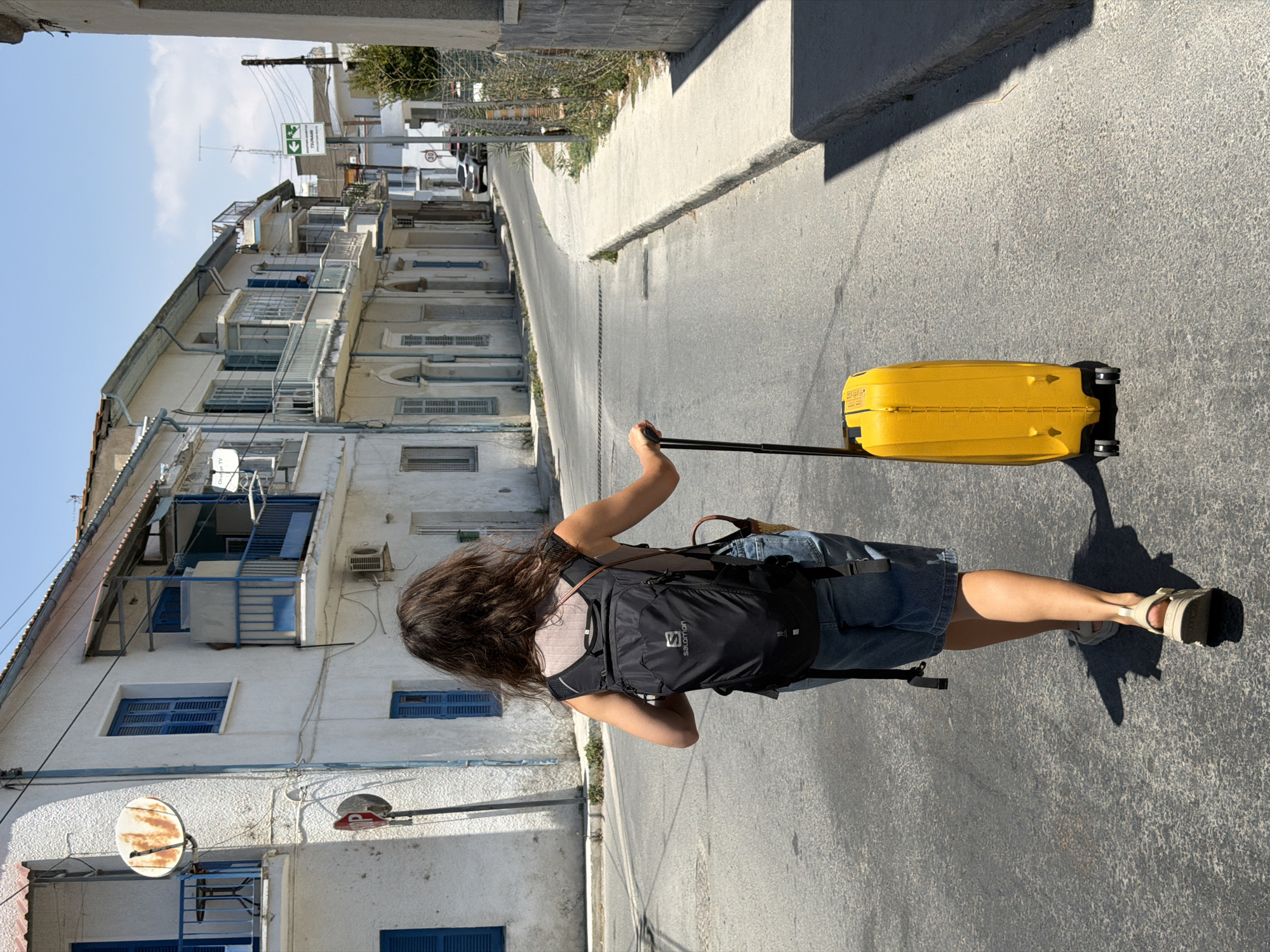 A person walks down a paved street in a European city. The person is seen from behind and wears a black backpack and denim shorts. They are pulling a bright yellow suitcase on wheels. The street is lined with white buildings with blue shutters and balconies. The sky is clear and blue, and the sun casts shadows on the ground. There are cars parked along the side of the street and a speed limit sign indicating 30.