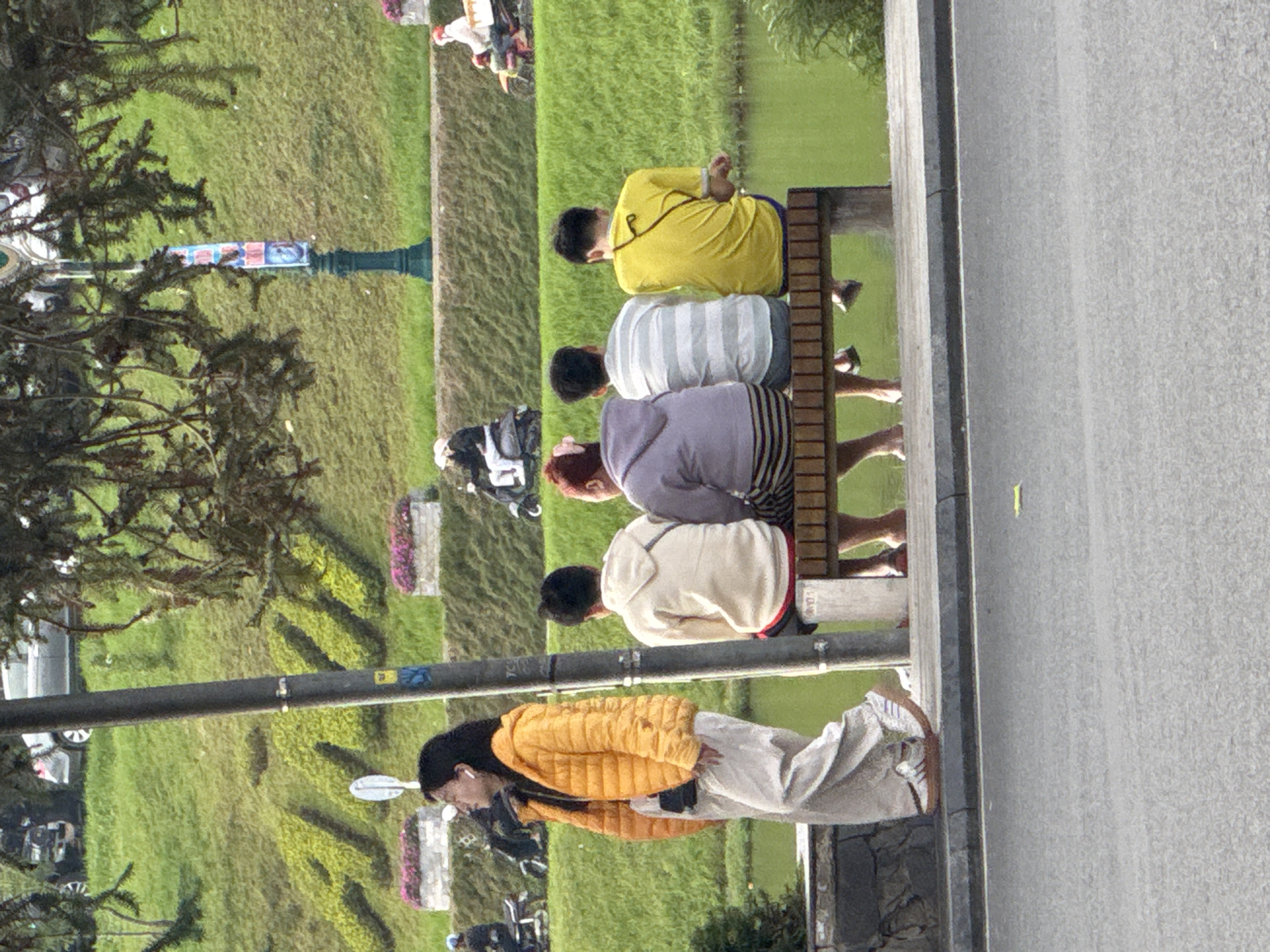 This detailed image depicts a daytime scene in Da Lat, Vietnam, encompassing an urban street, a park area with a water feature, and a sloped embankment. In the foreground, a grey asphalt road is separated by a dark grey curb from a paved walkway.
To the left of the frame, on the paved walkway, a woman with long dark hair walks towards the left. She is wearing a bright yellow puffer jacket, light beige wide-leg trousers, and white sneakers with brown soles. White earbuds are visible in both ears, and a black bag is worn across her torso.
Slightly right of the image center, a tall, dark grey utility pole stands vertically. To the right, a wooden bench accommodates four individuals, seated with their backs primarily towards the viewer, facing a calm, greenish-brown body of water. From left to right on the bench:
1. An individual with dark hair, wearing a light purple hooded sweatshirt and grey and white horizontally striped pants.
2. An individual with dark hair, wearing a white and grey horizontally striped short-sleeved shirt and dark blue pants. This person is barefoot.
3. An individual with dark hair, wearing a yellow short-sleeved t-shirt and dark shorts, with bare legs and feet dangling.
4. An individual with reddish-brown hair, wearing a light purple long-sleeved top and grey and white horizontally striped pants.
Behind the water, a significant grassy embankment slopes upwards. The word "DALAT" is prominently spelled out on this slope using lighter green foliage. Two rectangular white stone planters, each containing pink flowers, are positioned on the embankment. An ornate dark green lamppost adorned with a colorful vertical banner stands on the embankment towards the right. In the far background, on an elevated road, several vehicles including cars and motorcycles are visible; a person on a dark motorcycle is on the left, and a person on a white scooter is on the right. Dark green foliage from trees partially obscures the upper portion of the scene. The lighting indicates daylight conditions.