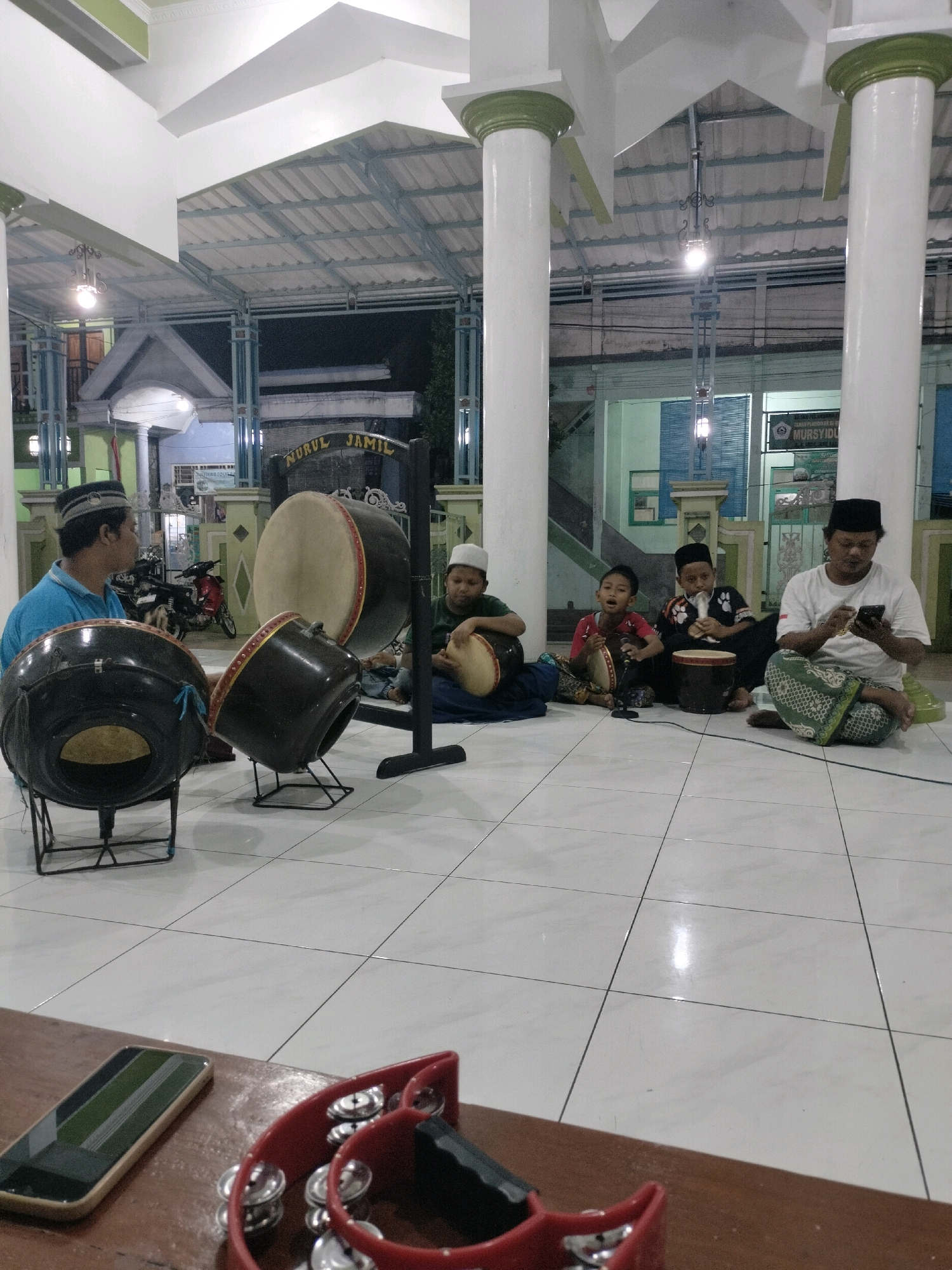 The image depicts a group of people, including men and children, gathered on a tiled floor, likely in front of a building with a corrugated metal roof supported by large white columns. The scene appears to be taking place at dusk or night, illuminated by artificial lighting. Several large drums, possibly rebana or kompangan, are set up on stands and on the ground. Some individuals are actively playing or preparing to play the drums. One man, wearing a blue shirt and a peci (a type of cap), is seated and looking towards the drums. A man in a white shirt and patterned sarong is seated to the right, looking at his smartphone. Two children are also present, one in a red shirt and the other in a black and white patterned shirt, both holding and playing small drums. The background shows a building with signs, one of which reads "NURUL JAMIL" and another which is partially visible with the text "MURS ..." which could be part of "MURSIDUL" or similar. A motorcycle is parked in the background. The presence of these drums and the setting suggests a religious or cultural gathering, possibly related to Islamic traditions. The location is identified as the city of Wedung, Indonesia. In the foreground, a smartphone and a red tambourine with jingles are placed on a wooden surface.