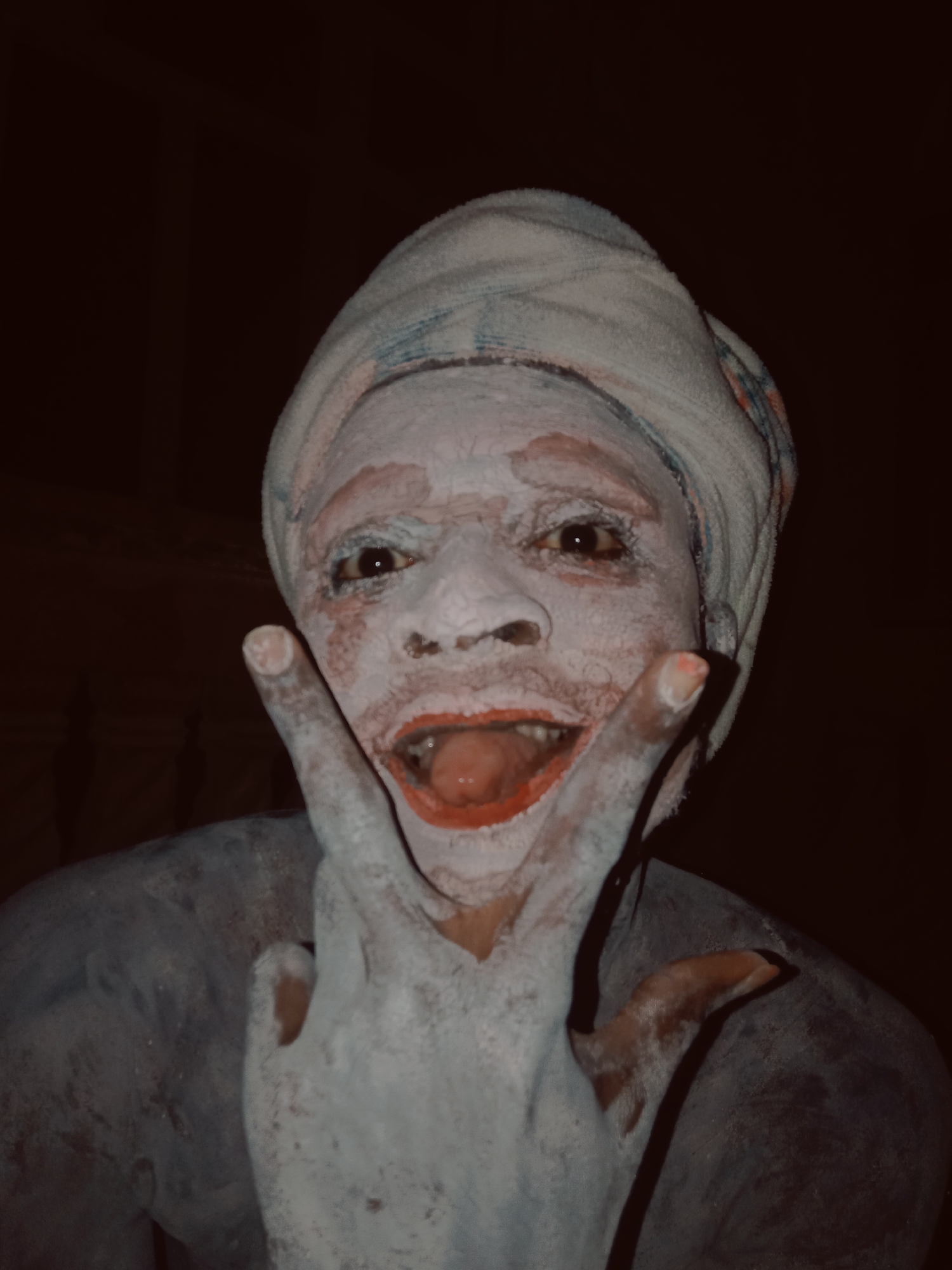 A person with white paint covering their face and body wears a towel on their head. The person is making a hand gesture in front of their face. Their mouth is open, and the inside of their mouth appears red. The image is dark, and the background is not visible. There is no verifiable location in Onitsha, Nigeria, indicated in the image.