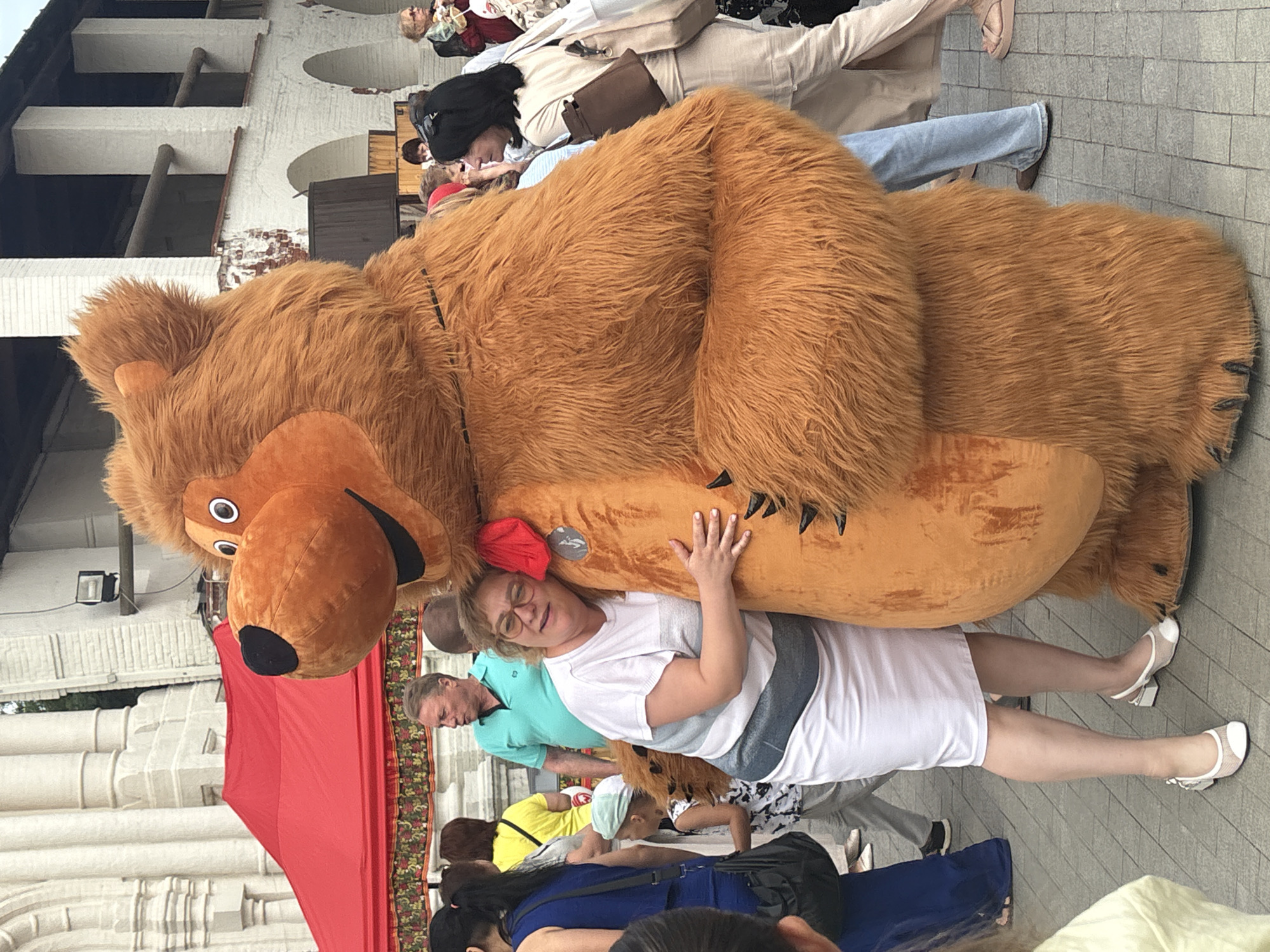 In Astrakhan, Russia, a large, fluffy brown bear mascot poses with a woman in a white dress and glasses. The woman is embracing the bear, with her right arm around its furry torso. She is wearing light-colored shoes with mesh detailing. Behind them, a red tent and a white brick building with arched windows are visible. Several other people are in the background, including a man in a teal shirt and a child in a patterned shirt and cap. The ground is paved with grey square tiles.