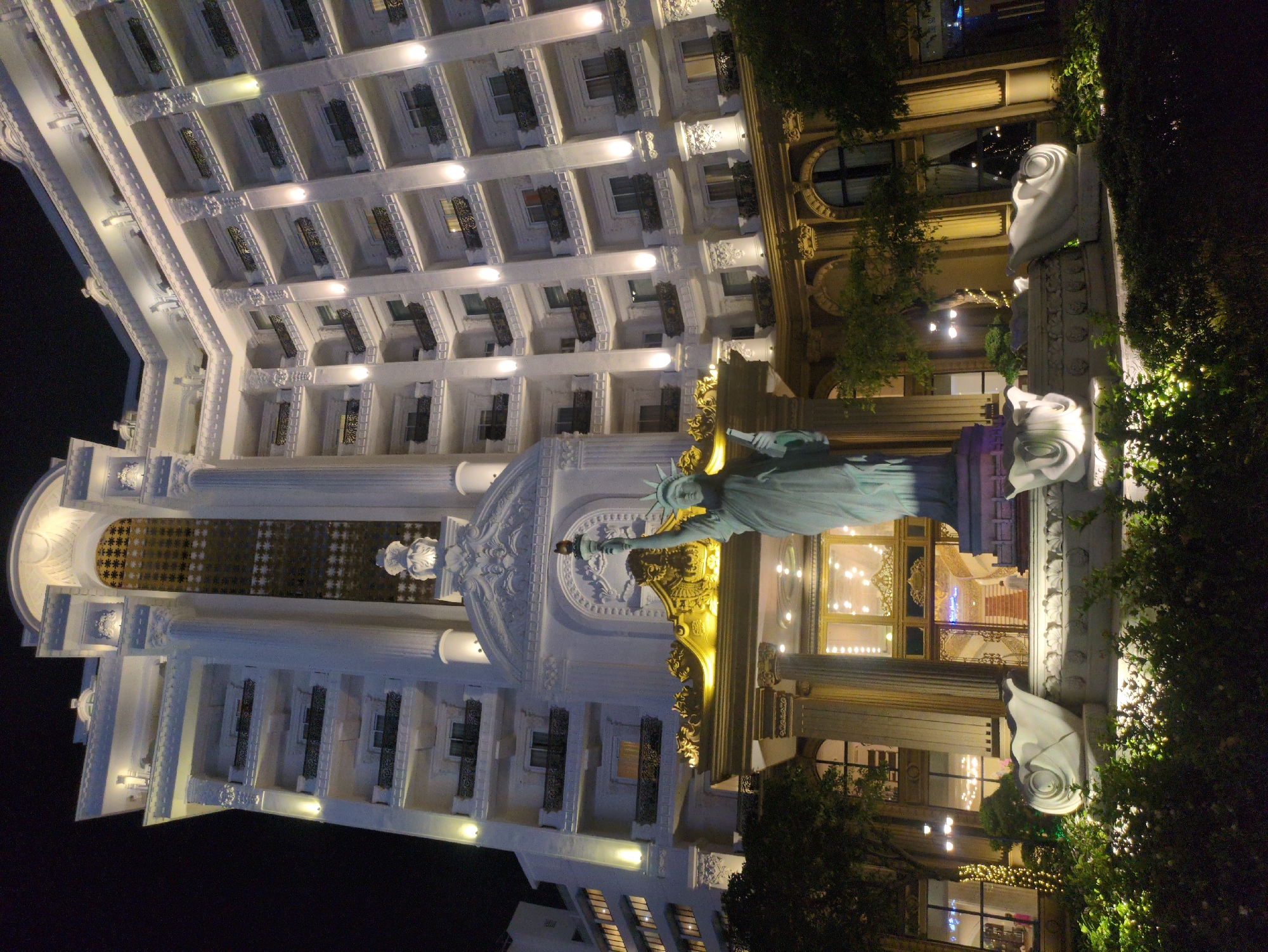 The image shows a building in Phu Quoc, Vietnam, featuring a replica of the Statue of Liberty. The building is brightly lit, with white and gold architectural elements. The Statue of Liberty is positioned in front of a building entrance with golden decorative elements. The facade is adorned with intricate details, including ornate window designs and classical columns. The surrounding area includes landscaping with trees and decorative white statues, enhancing the overall aesthetic of the location. The sky is dark, indicating that the picture was taken at night.