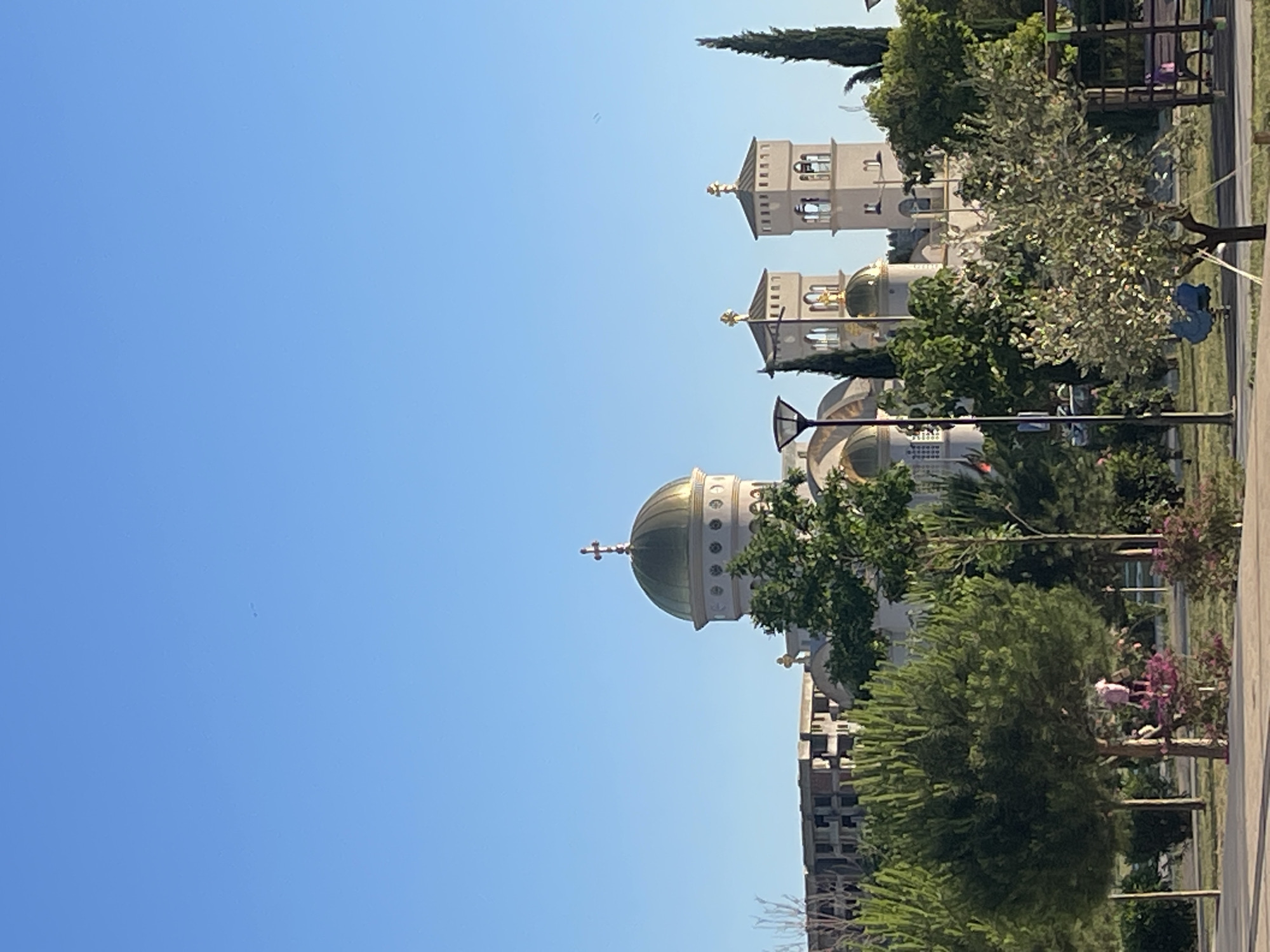 The image shows the Serbian Orthodox Church of St. Jovan Vladimir in Bar, Montenegro. The church complex includes a domed cathedral with a gold dome and cross, two bell towers, and various supporting structures. The church is surrounded by green trees and park land with walking paths. In the foreground, there is a person dressed in pink walking by a tree. In the background are low buildings. The sky is blue and clear.