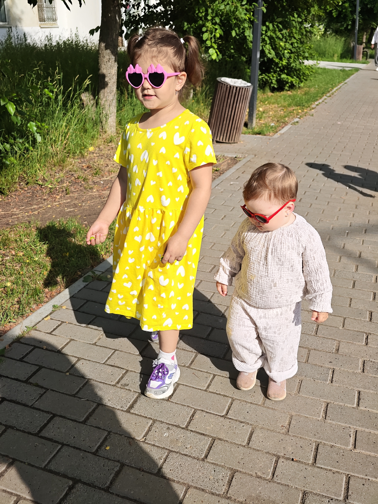 Two young children walk on a brick-paved sidewalk on a sunny day in Chaikovsky, Russia. The older child, on the left, wears a bright yellow dress with a white heart pattern and pink, heart-shaped sunglasses. They are also wearing white socks and purple and white sneakers. The younger child, on the right, wears a light-colored textured outfit and red sunglasses. A shadow of the older child is visible on the sidewalk. The background includes a grassy area, trees, and a trash can.