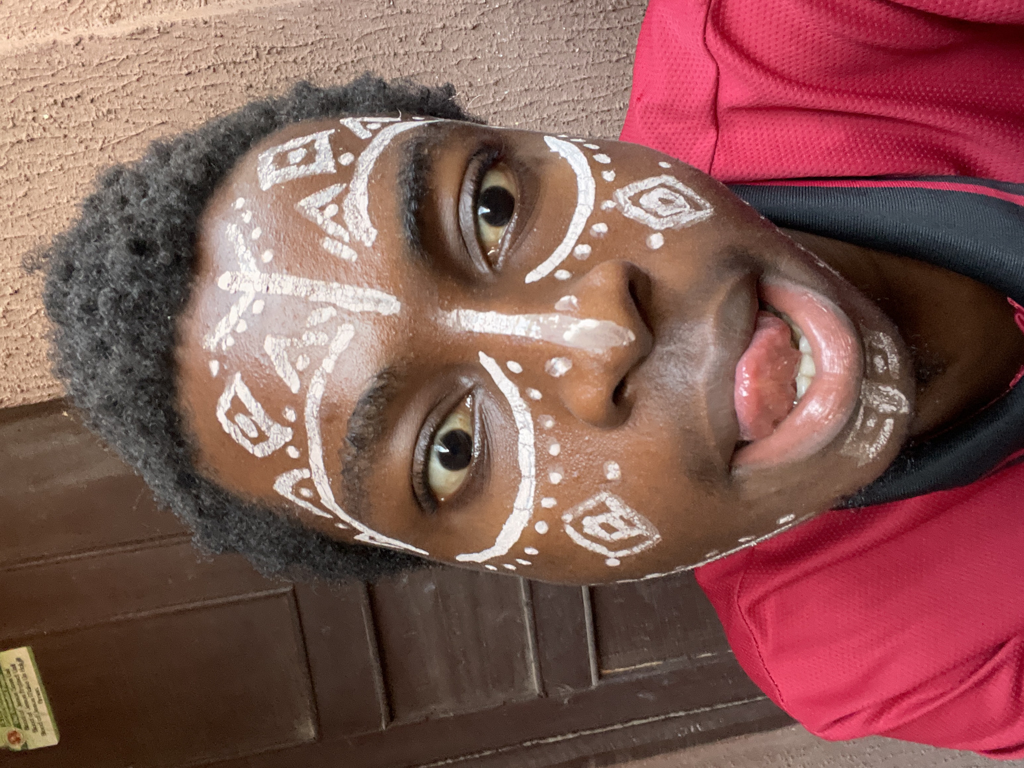 A close-up, rotated image shows a young, dark-skinned man with white paint on his face. The paint patterns include straight lines, dots, and geometric shapes. The man has short, curly hair and is wearing a red shirt. He is sticking his tongue out. The background consists of a brown, horizontal surface, possibly a wooden structure. The image was taken in Nkpor, Nigeria.