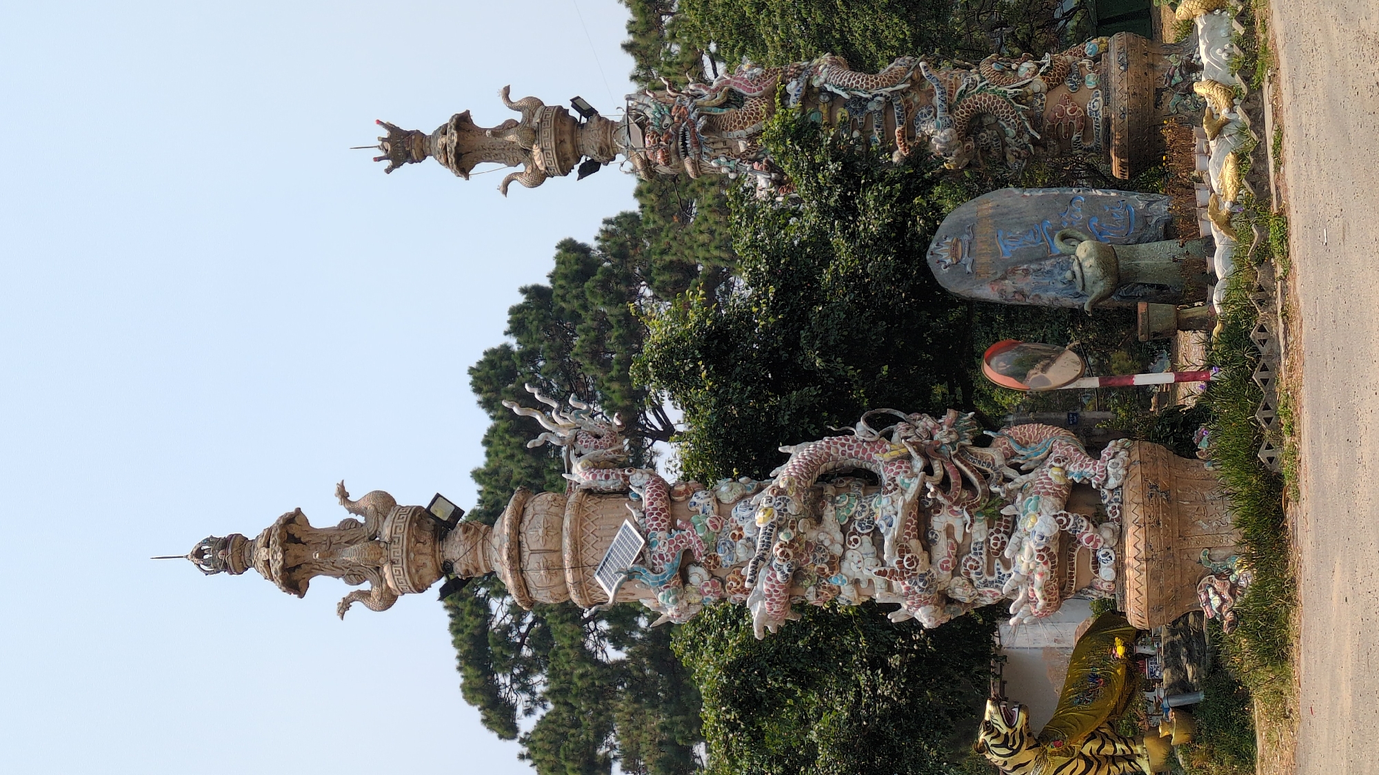 The image shows two ornate pillars in Lâm Sơn, Vietnam. The pillars are decorated with colorful dragon sculptures and topped with spire-like structures. A solar panel is attached to the side of one pillar. Lush greenery and trees surround the pillars, and a road is visible on the right side of the frame. Other statues and objects are visible in the background, including a large stone monument and a traffic mirror. The sky is pale blue.
