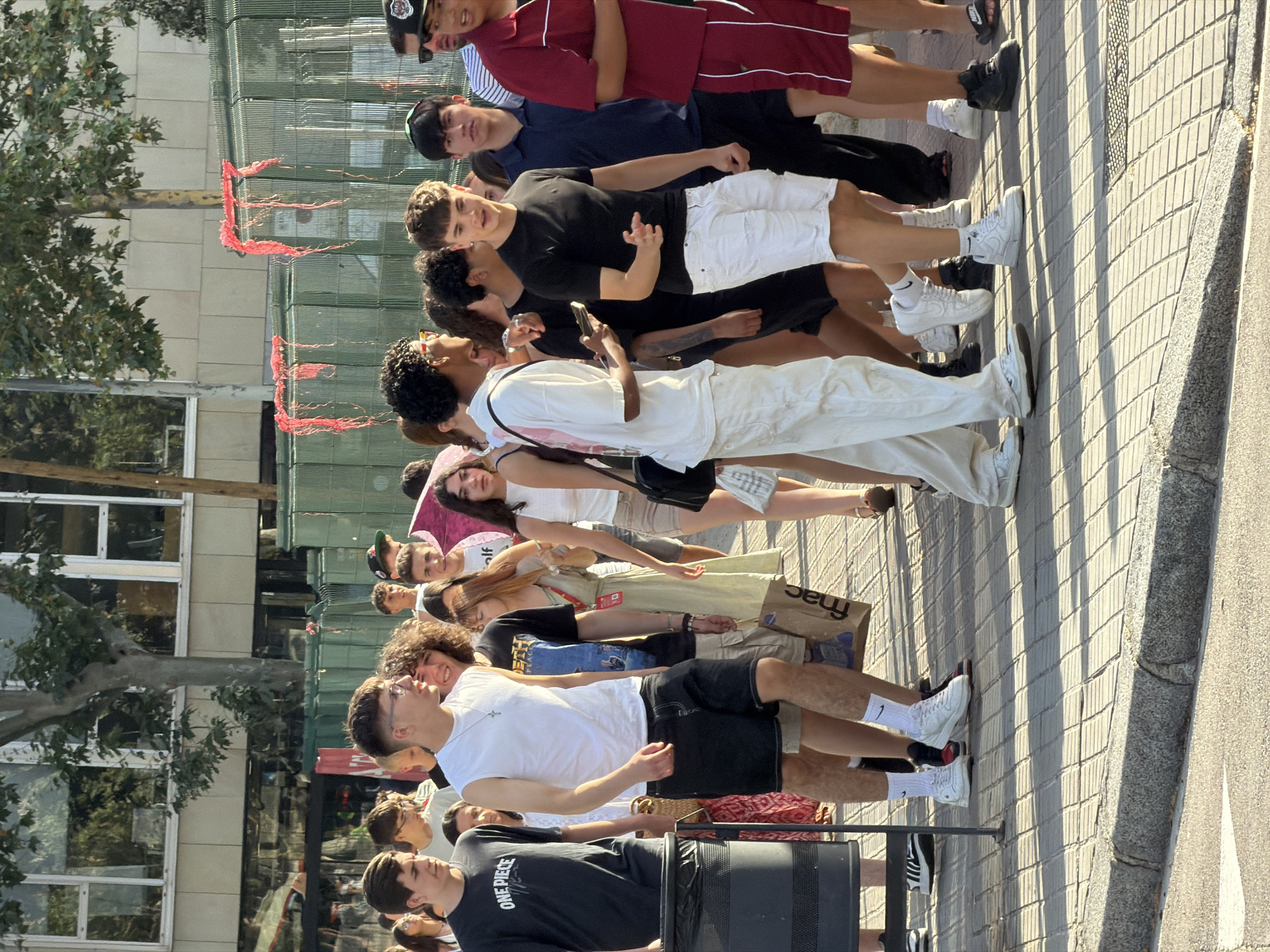 A group of young people walk along a city sidewalk in Barcelona, Spain. The sidewalk is paved with light-colored square tiles. In the background, a building with large windows and trees can be seen. A green metal fence with red streamers hangs over it separates the sidewalk from what appears to be an event or area beyond. The people are dressed in casual summer clothing, and some are carrying bags. The lighting suggests it is daytime, possibly late afternoon or early evening, with shadows cast on the sidewalk.
