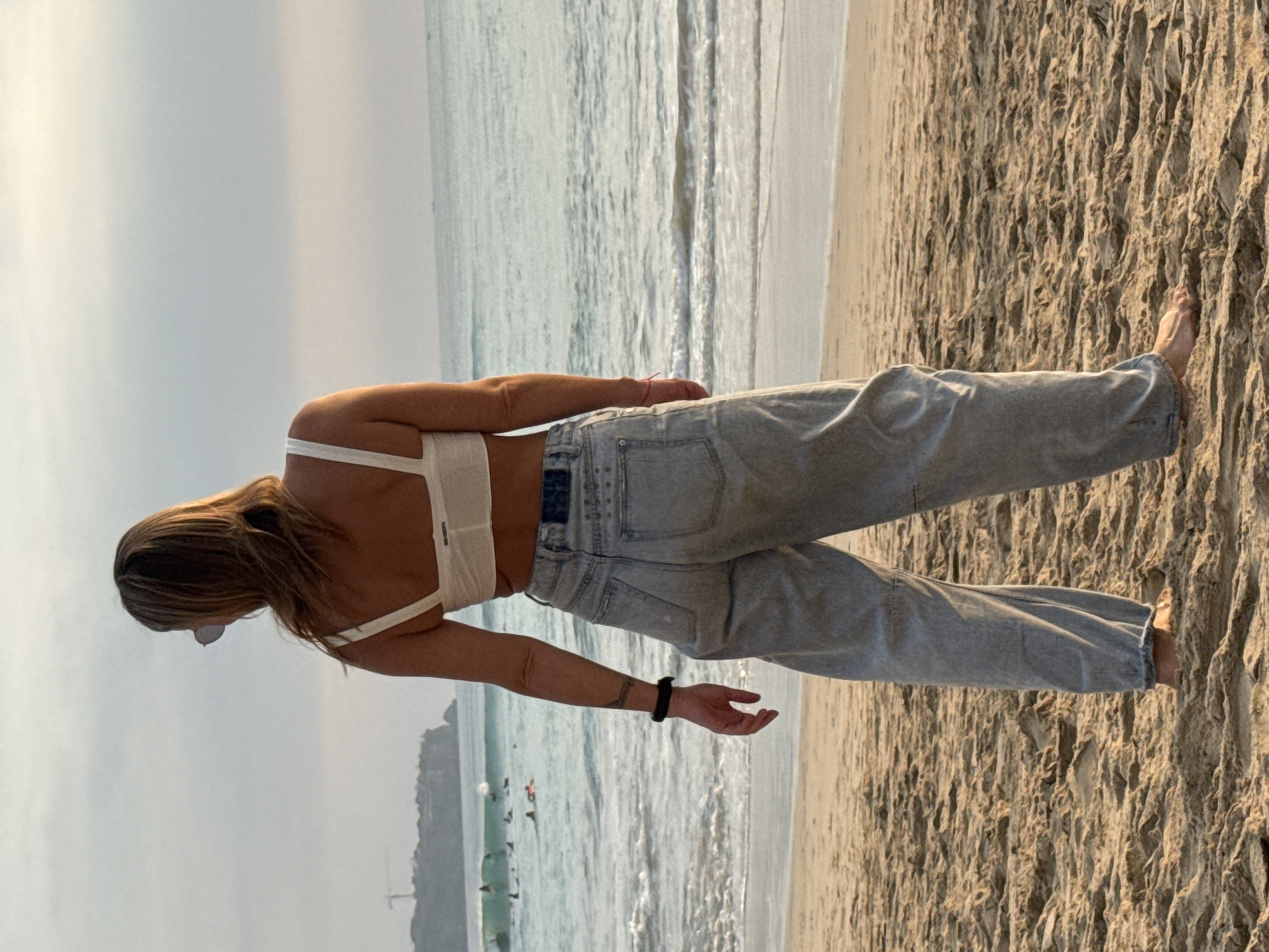 A person stands on a sandy beach in Kuta Selatan, Indonesia, facing the ocean. The person is wearing a white bikini top and loose-fitting light blue denim pants. They have blonde hair, wear sunglasses, and have tattoos on their left arm and hand. The ocean is calm with small waves, and several surfers are visible in the water. In the background, a cliffside with buildings and a construction crane can be seen. The sky is overcast with soft sunlight. The sand is textured with small dunes.