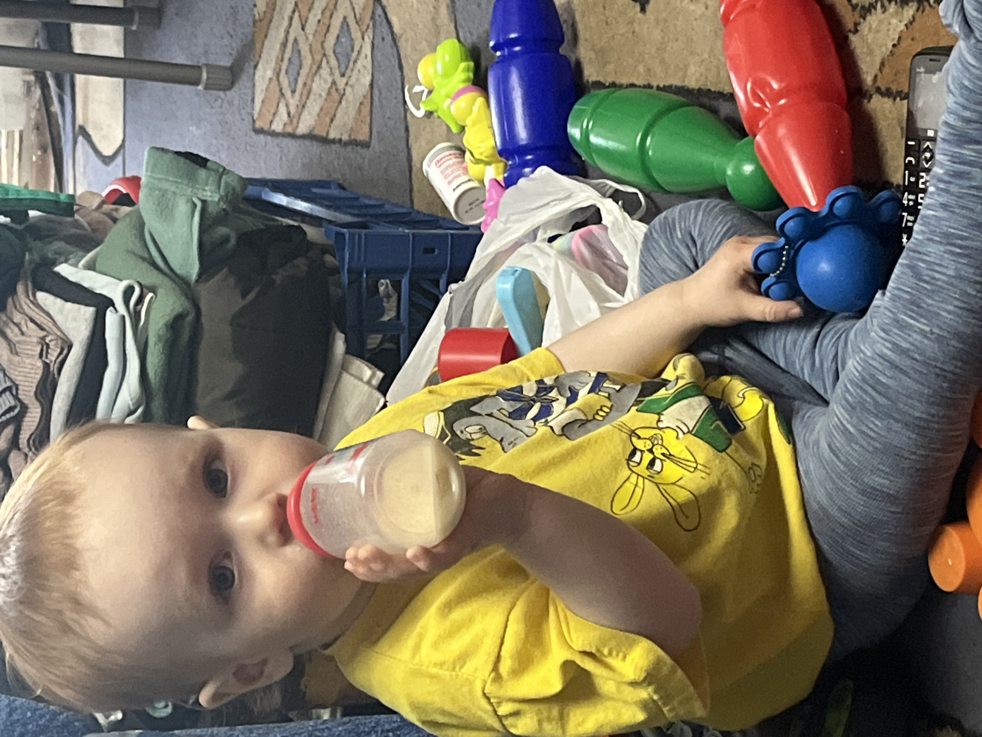A young child is lying on the floor with a baby bottle in their mouth and holding a blue toy in their hand. They are wearing a yellow T-shirt with cartoon characters and blue leggings. The floor is covered with a rug and cluttered with various plastic toys, clothing, and a blue crate. Based on the rug, the flooring material, and the presence of items typically found in a home, the location appears to be inside a residence. No evidence suggests a location in Chaikovsky, Russia.