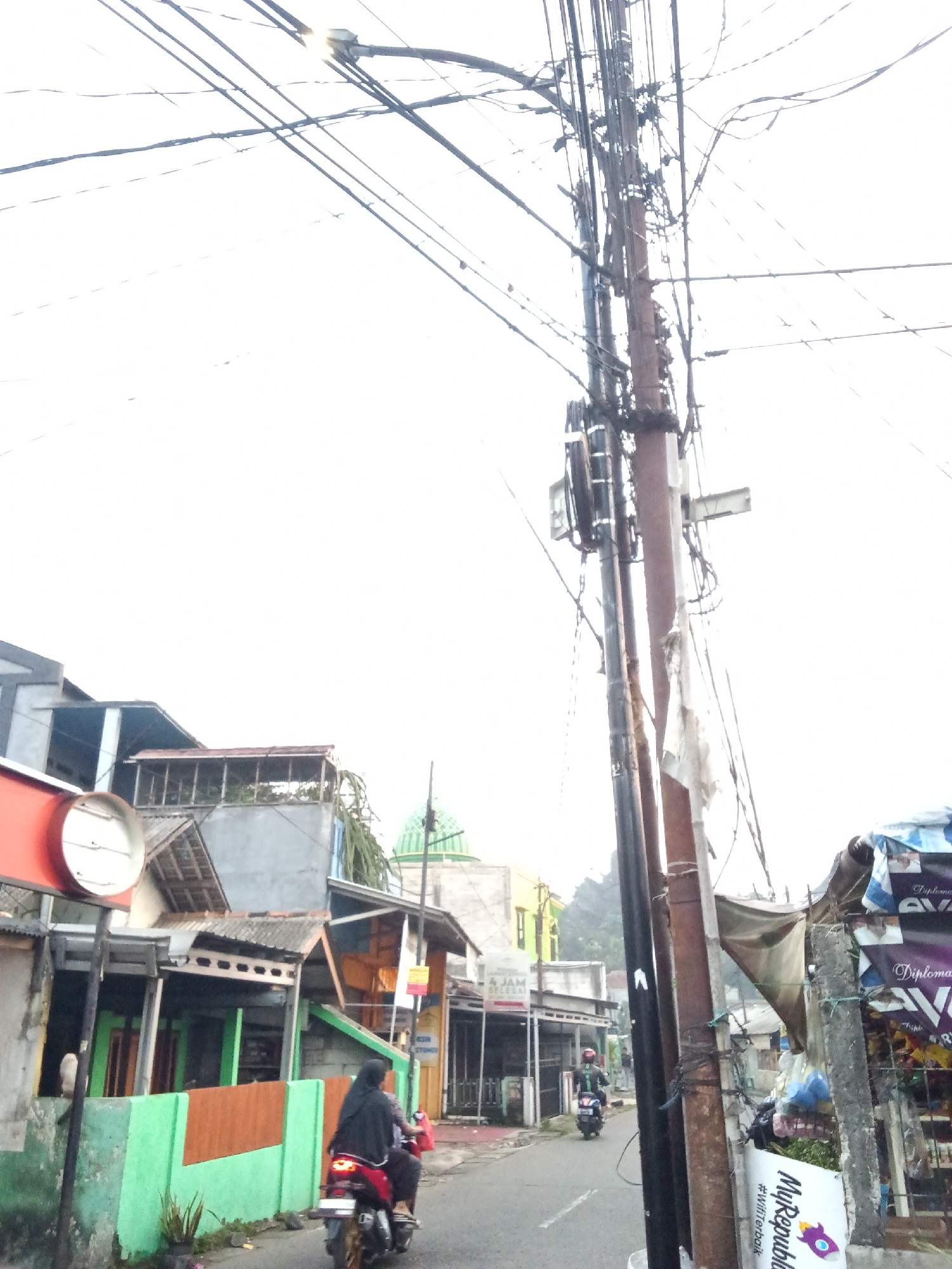 The image depicts a street scene in Depok, Indonesia, characterized by a dense network of overhead electrical wires and a utility pole dominating the center-right of the frame. The sky is overcast and bright. On the left side of the street, several buildings are visible, including a structure with an orange circular sign. A woman wearing a hijab is riding a motorcycle away from the camera down the asphalt road. Further down the road, another person on a motorcycle is visible. The buildings appear to be modest in design, with some having pitched roofs. A green dome, possibly part of a mosque, is visible in the background. The right side of the street shows more commercial establishments, including stalls with awnings and signage, one of which displays the text "MyRepublic" and a rocket graphic. The overall impression is of a busy, somewhat cluttered urban environment.