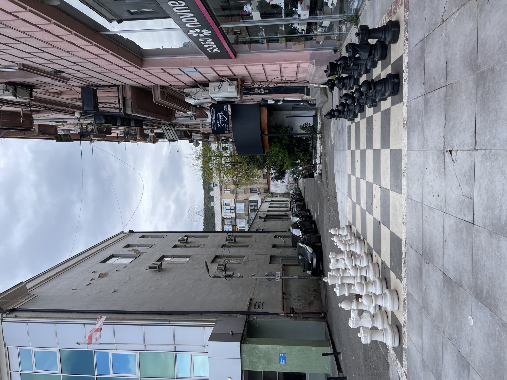 The image shows a street scene in Tbilisi, Georgia. A large-scale chessboard is painted on the ground, with oversized black and white chess pieces arranged at opposite ends. Cars are parked along the side of the street. A building with a blue glass facade and a Georgian flag is visible on the left. Across the street are shops, cafes, and apartments. In the background is the Tbilisi Ferris Wheel. The sky is overcast.