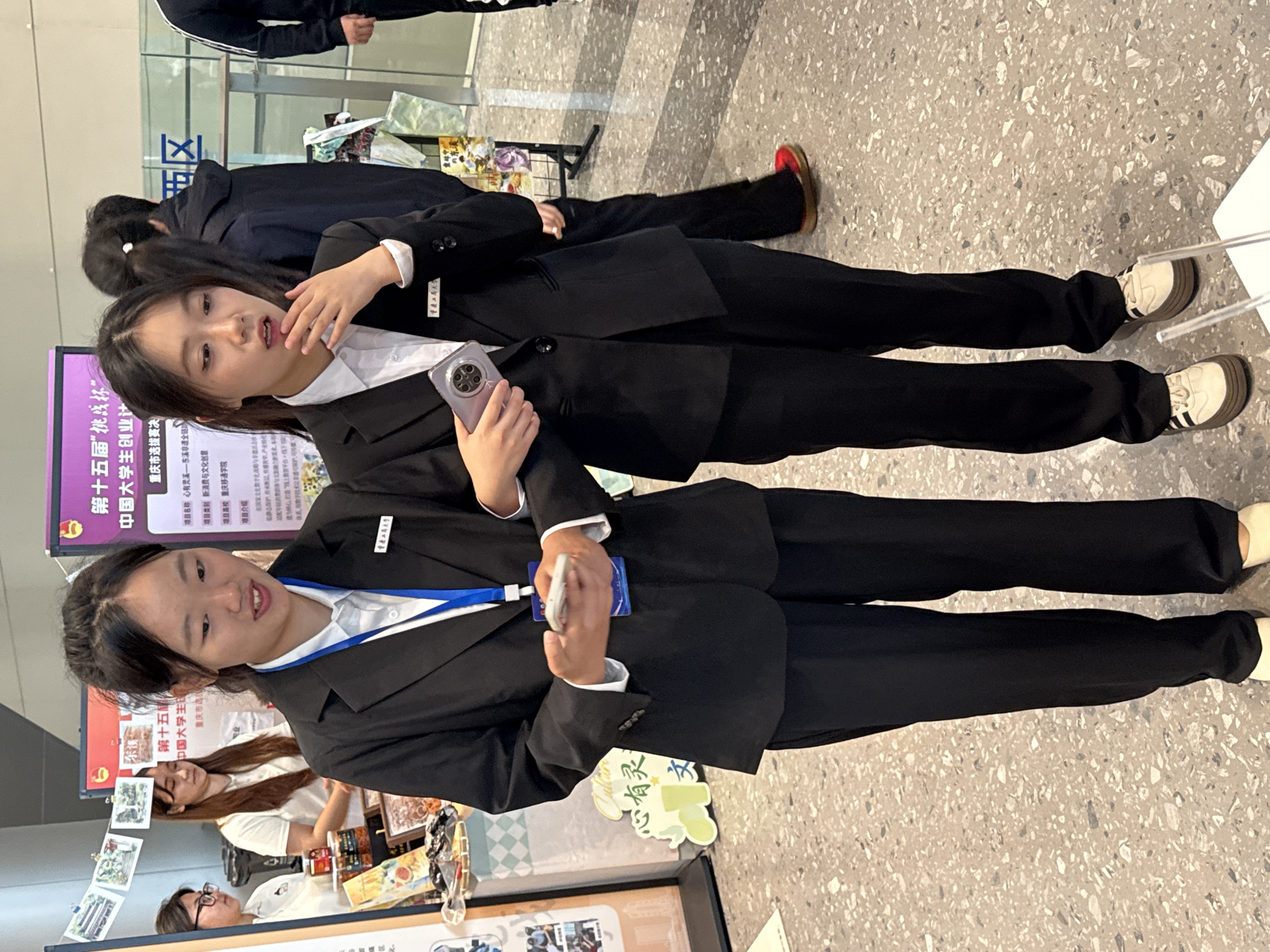 Two young women in black blazers and pants, wearing white shirts and lanyards, stand facing forward. The woman on the left holds a smartphone in her left hand and appears to be showing it to the woman on the right, who is touching her chin with her right hand and looking at the phone with an attentive expression. They are positioned in front of a display booth with posters and products. A large poster with Chinese text and images is visible behind them, and on the right side of the frame, another poster with "西区" (Western District) is mounted on a wall. The floor is a speckled terrazzo. The setting is likely an exhibition or event, possibly in the Chongqing High-tech Zone, China, given the context provided by the "西区" sign.