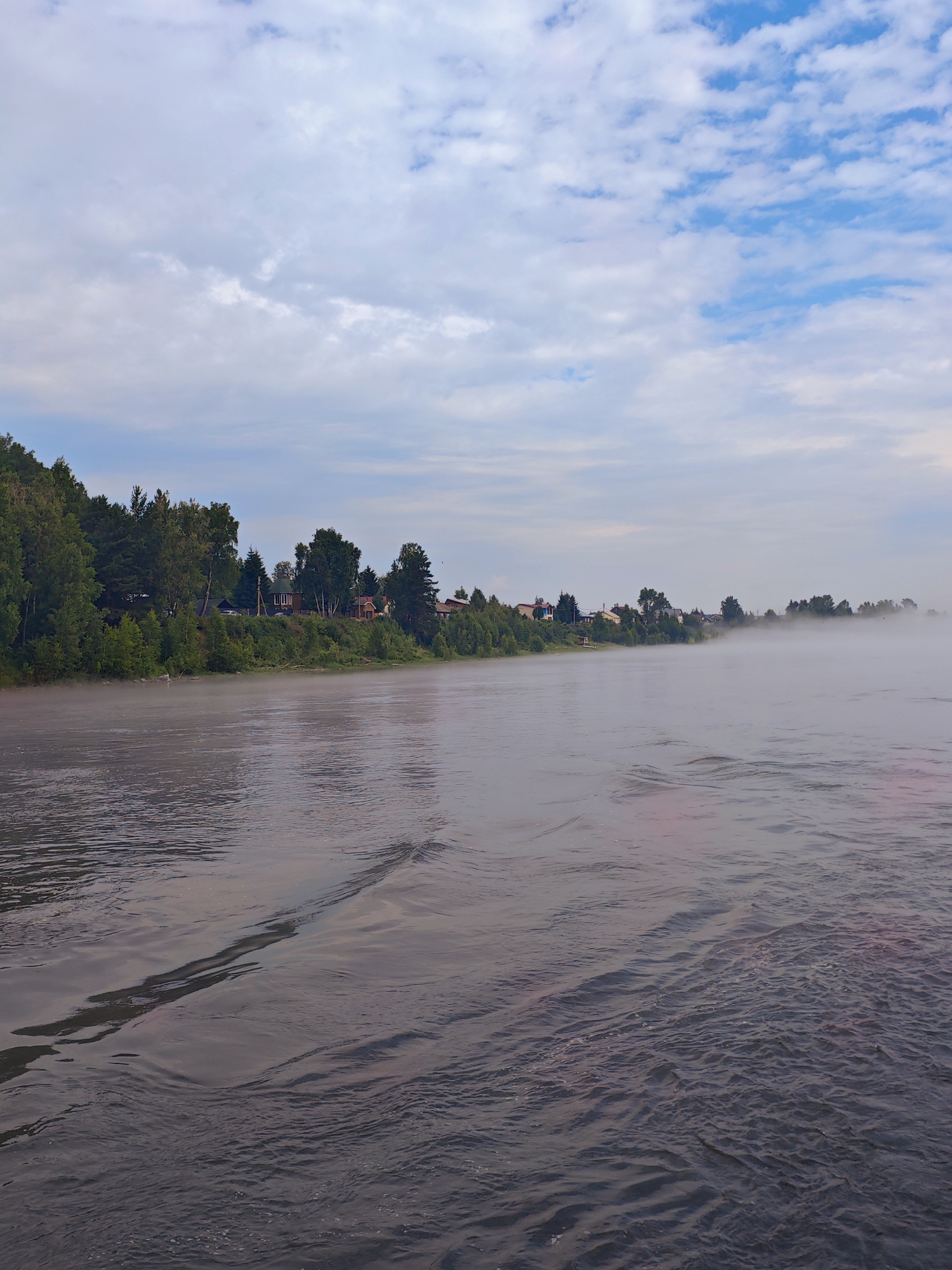 An overcast sky with scattered white clouds over a wide, calm river. A bank of trees and houses lines the left side of the river, with a dense forest in the background. A light fog or mist hovers over the water in the distance. The foreground shows ripples and waves on the river's surface. The scene depicts a quiet, rural landscape.