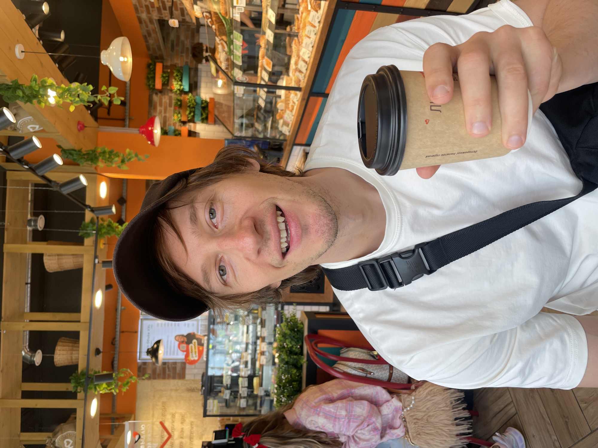A man wearing a white t-shirt and a dark baseball cap holds a coffee cup in his right hand, angled towards the camera. He is smiling and looking directly at the camera. The man is seated at a table in a bakery, with a display case filled with baked goods visible in the background. To the left of the man, a woman wearing a pink checkered shirt with red bows in her hair is seated at another table. The bakery has wooden beams on the ceiling, hanging plants, and orange and brick walls. The overall impression is a casual moment captured within a bustling bakery.