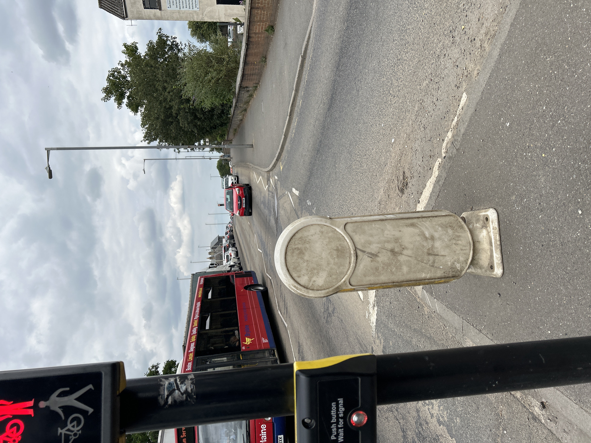 This media file captures a street scene in Boston, United Kingdom, with a focus on a pedestrian crossing. A red bus, with "Boston Lincoln Skegness Spalding Sleaford Boston" visible on its side, is prominent in the mid-ground, suggesting it is a local service. Several cars are also visible in traffic, indicating moderate road activity. The foreground features a pedestrian signal button with the text "Push button Wait for signal," and a red light indicates the signal is active.
The scene appears to be set during the day, under a cloudy sky. The presence of the pedestrian crossing, street lights, and road markings firmly place the setting within an urban or suburban environment. The overall mood is neutral, depicting a typical moment of daily urban transit. The image is oriented vertically.
