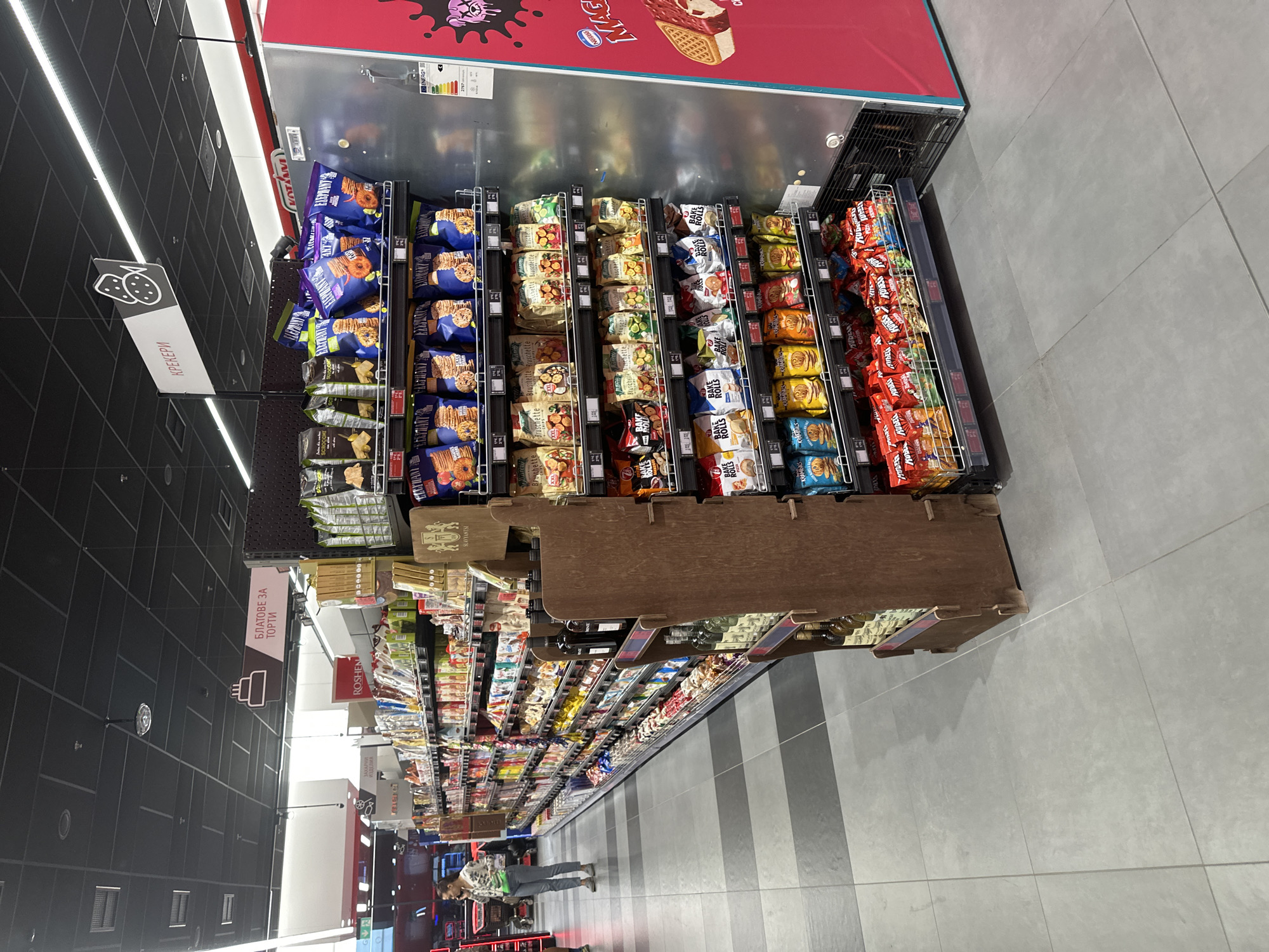 This image captures a long aisle within a well-lit supermarket or grocery store in Sofia, Bulgaria, viewed from one end looking down its length. The scene is dominated by shelves heavily stocked with various packaged food items.
On the immediate left (upper part of the rotated image), a refrigerated display unit features a red top border and an energy efficiency label. Above this unit, a vibrant graphic shows an ice cream sandwich with the "NESTLE" logo and text "merci". Below this unit, multiple rows of packaged goods, primarily crackers and cookies, are displayed on metal racks. Notable brands include "ELEPHANT," "KRAMBALS," "Bruschette," and "BAKE ROLLS." These shelves extend further into the distance, showcasing a variety of sweets, snacks, and baking ingredients.
Hanging from the dark, grid-patterned ceiling are white rectangular signs with Cyrillic text. One prominent sign reads "КРЕКЕРИ" (Crackers) with a cracker icon, indicating the section's contents. Further down the aisle, another sign reads "БЛАТОВЕ ЗА ТОРТИ" (Cake Bases) with a cake icon.
The floor is composed of large grey tiles, with alternating lighter and darker striped sections running perpendicular to the aisle. In the distance, a person is visible browsing items, indicating active shopping in the store. Other visible texts on products include "ROSHEN," "SLAVYANTSI," and "KUDE KUBE." The overall impression is that of a modern, organized retail space.