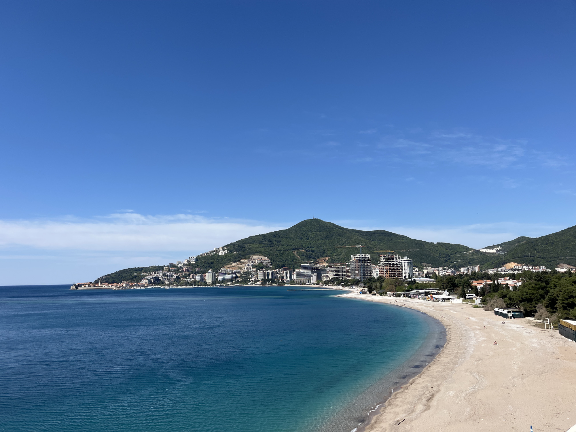 The image depicts a coastal scene in an urban setting. In the foreground, a sandy beach curves along the coastline, bordered by clear blue water. Scattered people can be seen on the beach.
Behind the beach, a dense collection of buildings and structures extends up a hillside towards a prominent mountain. The buildings are of various heights and styles, indicating a mix of residential and commercial developments.
The sky is bright blue with scattered clouds. The overall scene suggests a sunny day at a resort town or city situated on the coast. The location is Budva, Montenegro.