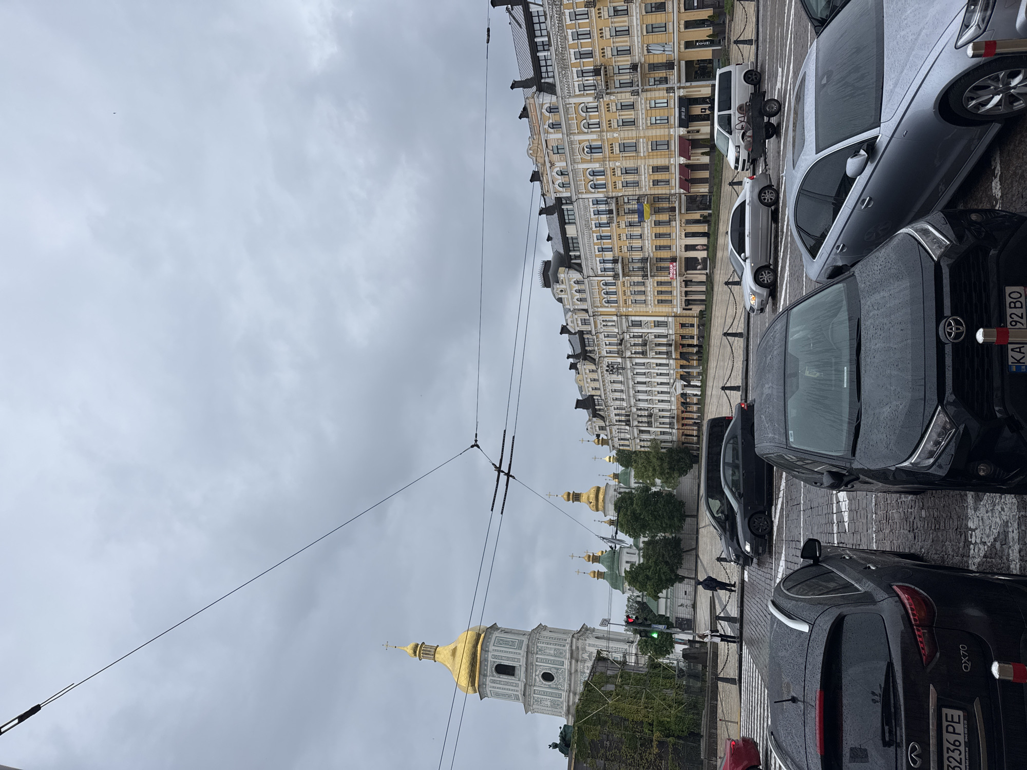 The image portrays an overcast daytime street scene in Kyiv, Ukraine. The prominent landmark on the left is the magnificent St. Sophia's Cathedral, identifiable by its distinctive golden domes and a tall white bell tower. Several other golden-domed structures are visible behind it, partially obscured by trees. On the right, a long, multi-story building with an ornate yellow facade and numerous dark windows lines the street, with a small Ukrainian flag hanging from one of its balconies.
The foreground and mid-ground are filled with a street made of cobblestones, featuring a zebra crossing. Numerous cars are parked along the street and in traffic lanes; their wet surfaces suggest recent rain. A black Toyota SUV displays the Ukrainian license plate "KA 92 BO," while a black Infiniti QX70 (model "QX70") shows "3236 PE" on its rear license plate. A silver Audi sedan and a white van pulling a small trailer are also visible. Overlapping power or tram lines traverse the grey, cloudy sky.
A few indistinct pedestrian figures can be seen on the sidewalk in the distance, near the cathedral, and traffic lights are visible, one showing green. The scene depicts a typical, quiet day in the city, characterized by its historic architecture and urban infrastructure under a uniformly grey sky.