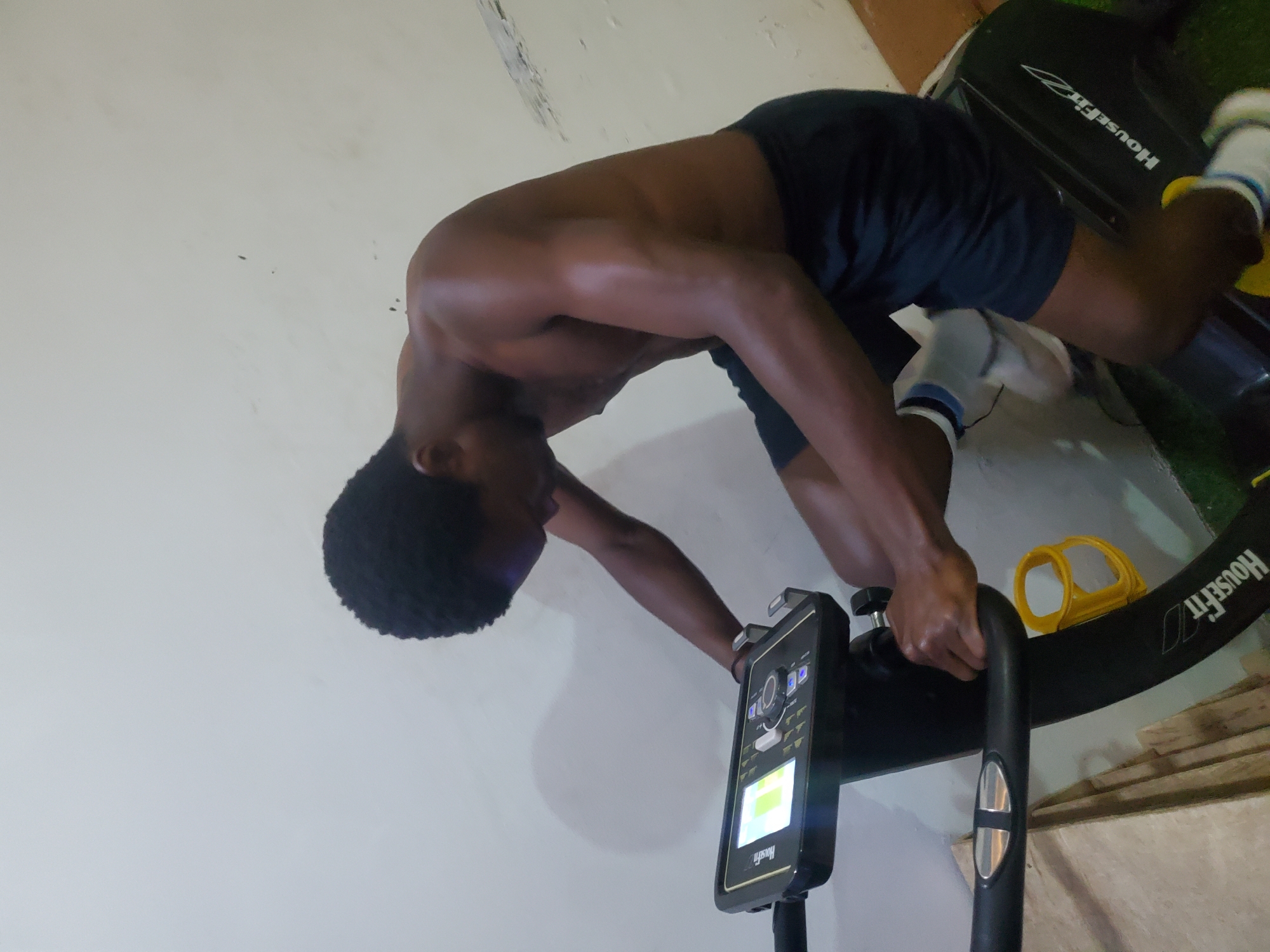 A shirtless Black man with dark skin and short black hair is exercising on a treadmill. He is leaning forward over the console of the treadmill, his hands gripping the handles. His body is muscular, and he appears to be sweating. He is wearing black shorts and white socks with blue and black stripes. The treadmill is black with a yellow handle and a digital display. The background is a plain white wall. There is a logo on the treadmill that says "HouseFit". The lighting suggests it could be indoors, and the time of day is not discernible. The scene is set in Jos, Nigeria. The man's posture and exertion suggest he is engaged in an intense workout. The overall mood is one of fitness and determination.