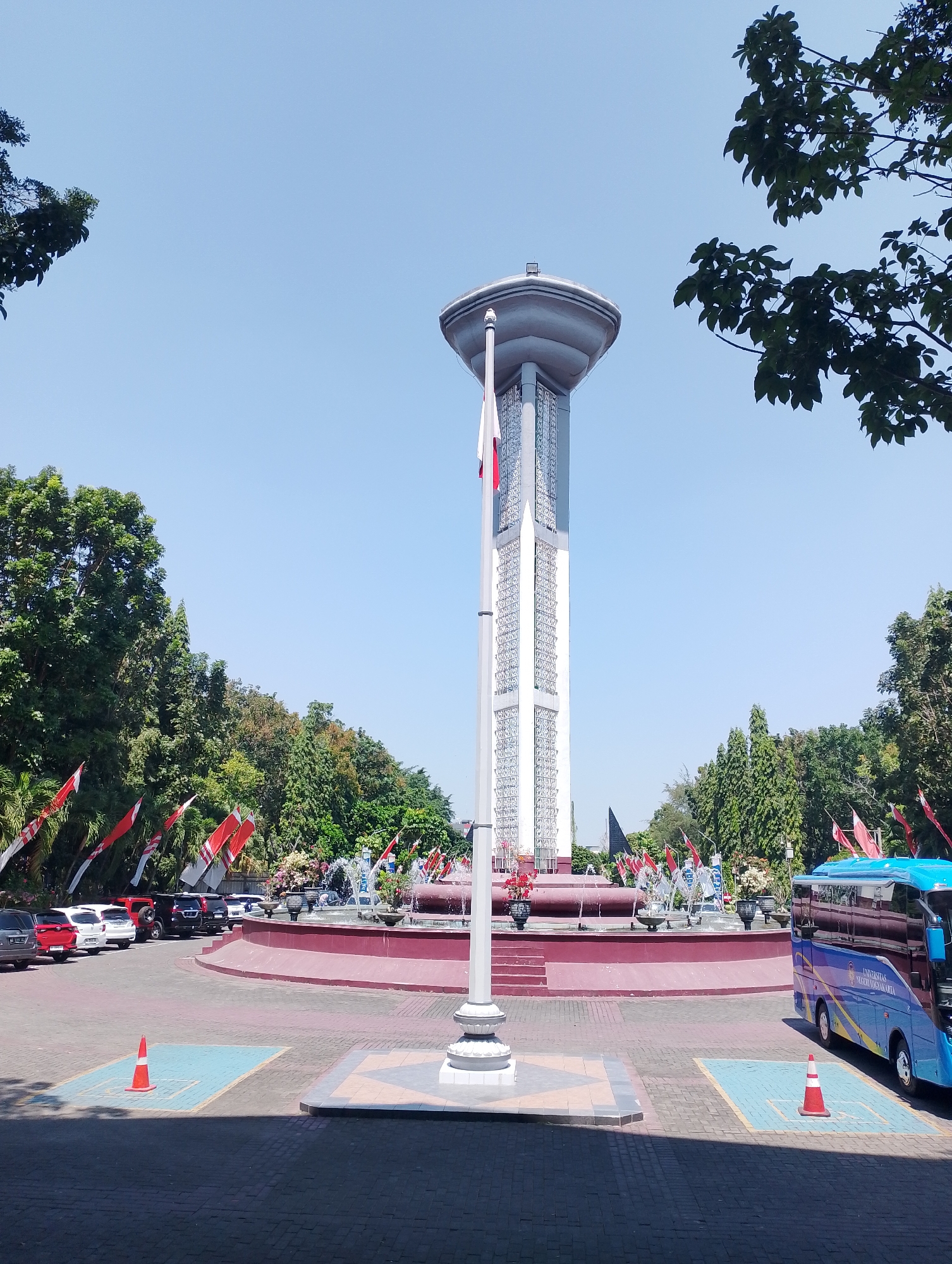 The image depicts the Tugu Lonceng (Bell Tower) monument in Depok, Indonesia, on a clear, sunny day. The monument, a tall, slender structure with a distinctive bell-shaped top, stands prominently in the center of a circular plaza. The tower is adorned with intricate patterns on its sides and features a flagpole flying a red and white Indonesian flag. Surrounding the base of the tower is a multi-tiered fountain with water jets, decorated with various red and white flags lining the perimeter of the fountain area.
To the left of the monument, a parking lot is visible, filled with several cars of various colors, including red, white, and black SUVs and sedans. Orange traffic cones are placed on blue-marked parking spots. Beyond the parking lot, dense green trees form a lush backdrop, with some trees exhibiting autumnal colors.
To the right of the monument, a blue bus with yellow accents and text reading "UNIVERSITAS NEGERI YOGYAKARTA" is parked. Another orange traffic cone is visible on a blue-marked parking spot in front of the bus. The ground in the foreground is paved with dark brick pavers, with lighter colored paving stones forming geometric patterns. The overall scene suggests a public space, possibly a tourist attraction or a significant landmark within the city of Depok. The presence of numerous flags indicates a festive or patriotic occasion.
