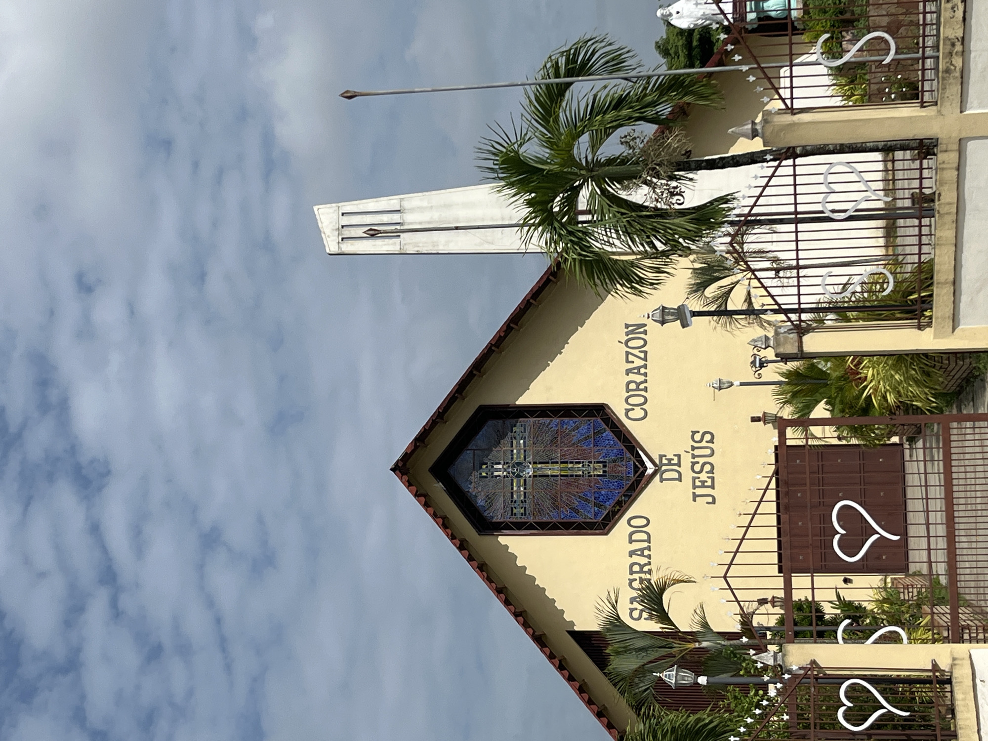 The image shows the facade of a church, likely in Tinaquillo, Venezuela, with the inscription "SAGRADO CORAZON DE JESUS" visible on its pale yellow exterior wall. A large, hexagonal stained-glass window depicting a cross is centrally placed on the upper part of the facade. To the right of the church, a tall, slender white bell tower rises against a cloudy sky. Palm trees and other greenery are visible in the foreground, obscuring parts of the entrance gate and fence. The gate is made of dark metal with decorative white heart shapes. A statue of a white figure, possibly a religious icon, is partially visible on the far right of the image. The scene is illuminated by daylight, suggesting an outdoor setting.