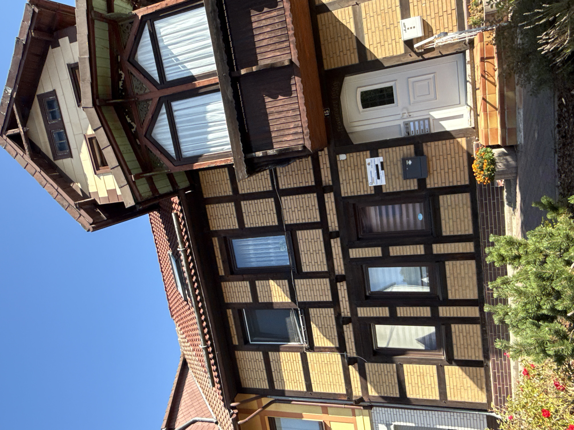 The image shows the exterior of a multi-story building in Bad Sachsa, Germany, with a clear blue sky overhead. The building features a distinctive half-timbered facade with dark wooden beams against yellow brickwork. The roof is covered with red tiles, and there is a prominent balcony with a wooden railing on the upper level. Several windows are visible, some with white curtains. In front of the building, there is a set of steps leading to a white door with a small window. A mailbox is attached to the wall next to the door. To the left of the entrance, a sign is affixed to the wall. In the foreground, there are various plants, including a small evergreen shrub and flowering bushes with red and orange blooms. The architectural style suggests a traditional German construction.