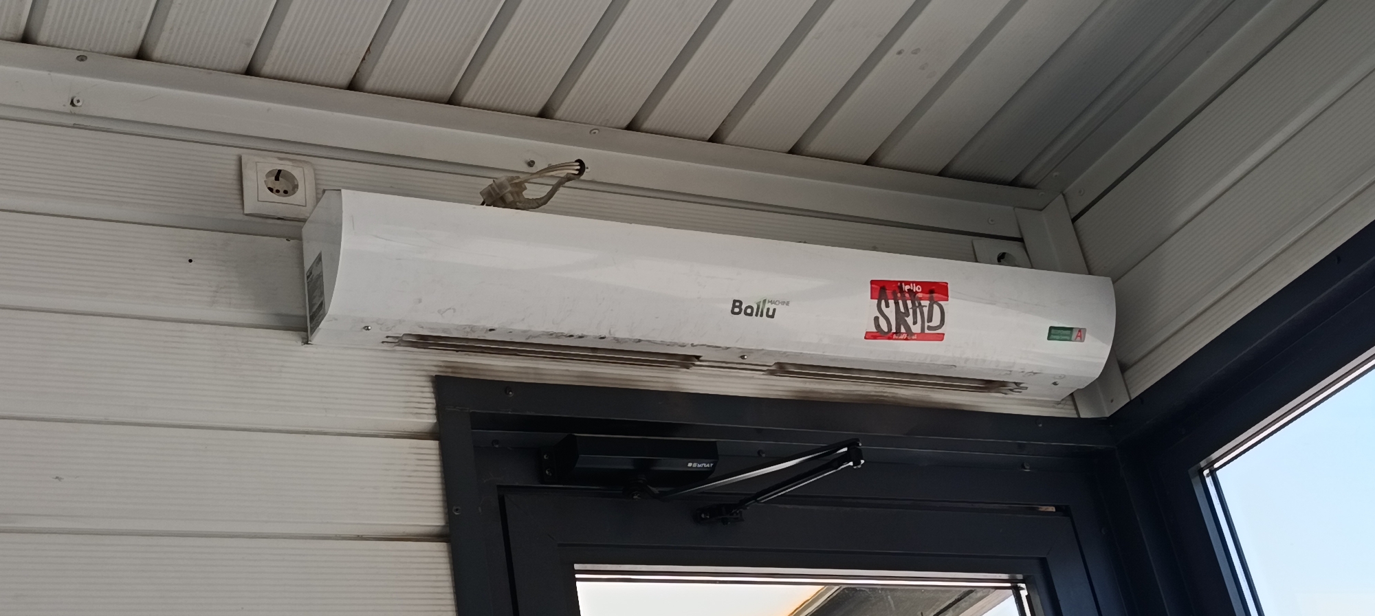 A white air conditioning unit is mounted on the exterior wall of a building, positioned above a dark-framed window or door. The building's exterior is clad in horizontal white siding. The ceiling above the air conditioner is made of ribbed white panels. To the left of the air conditioner, a white electrical outlet is visible on the wall. Wires extend from the top of the air conditioner unit. A red sticker with white text is affixed to the front of the air conditioner, partially obscuring the brand name "Ballu". Below the air conditioner, a door closer mechanism is attached to the frame of the window or door. The scene appears to be outdoors, with a clear blue sky visible through the window or door on the right side of the image. The location is identified as the city administration of Qarağandı, Kazakhstan.
