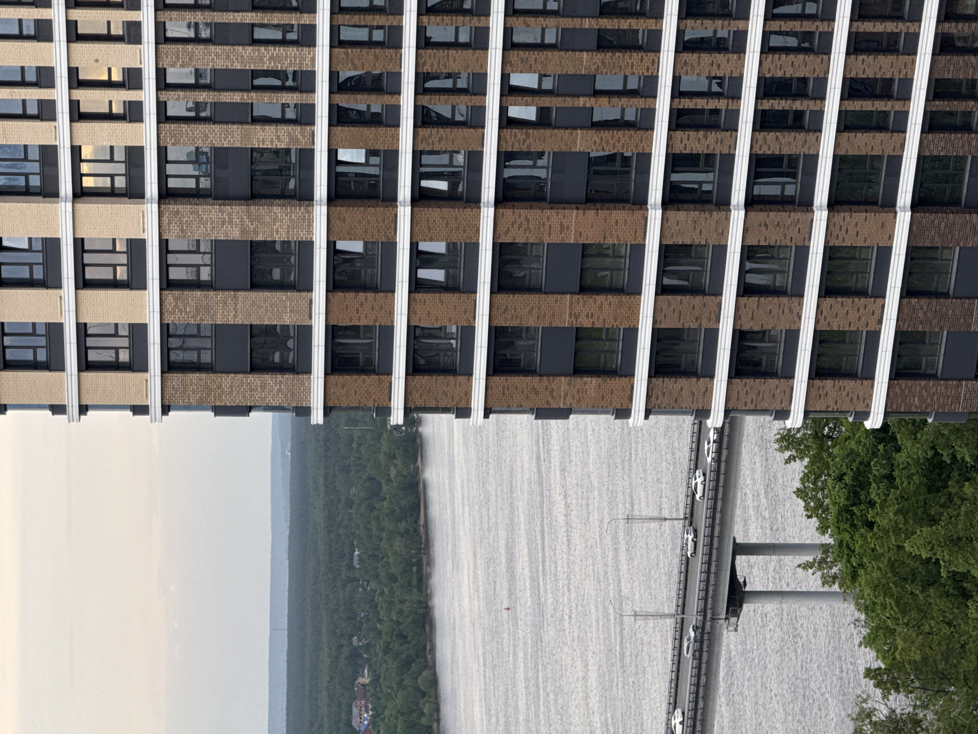 The image depicts a view of Perm, Russia, including a portion of a tall building with a repetitive pattern of windows and vertical white columns. Below the building, there is a body of water (likely the Kama River) with a bridge carrying vehicle traffic. Trees are visible on one side of the water. The sky appears hazy.