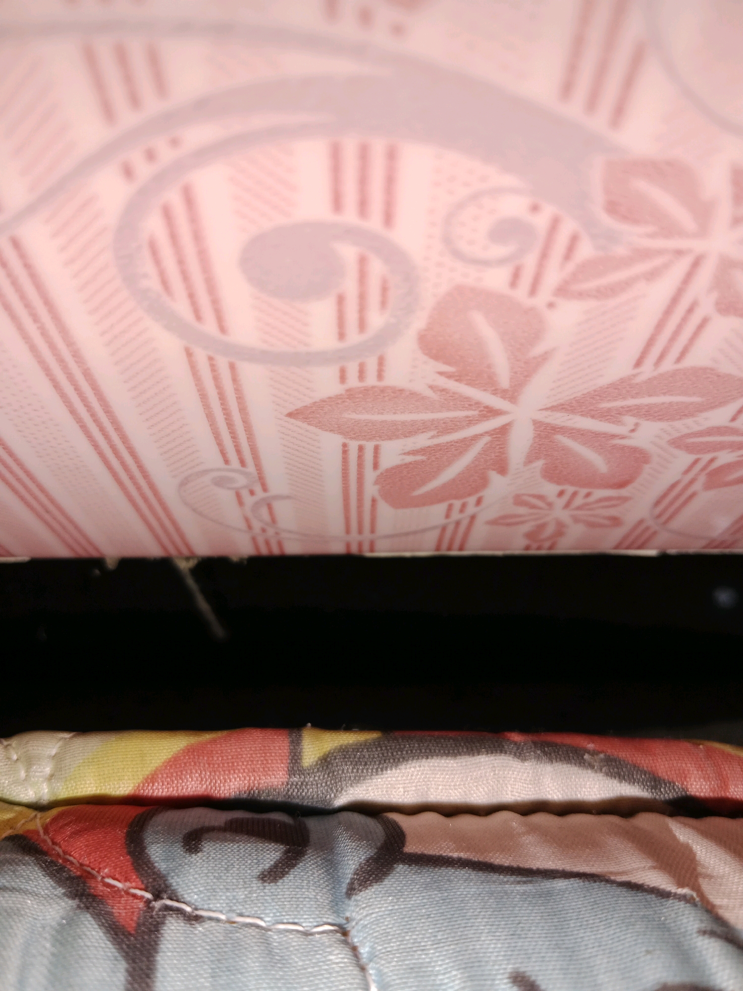 The image shows a close-up, low-angle view of layered fabric objects, likely bedding or cushions, in a dimly lit indoor setting. The uppermost layer features a pale pink fabric with an embossed pattern of stylized flowers and swirling lines, over a background of thin vertical pink lines. Below this is a black void, followed by two more layers of patterned fabric. The middle layer has a colorful abstract pattern with yellow, red, and black elements. The bottom layer is light blue with black graphic shapes. The fabrics are quilted, indicated by visible stitching. The image appears to be taken in a residential interior in Ringinrejo, Indonesia, likely a bedroom or living area. There are no people or overt actions depicted; the focus is solely on the texture and pattern of the materials.