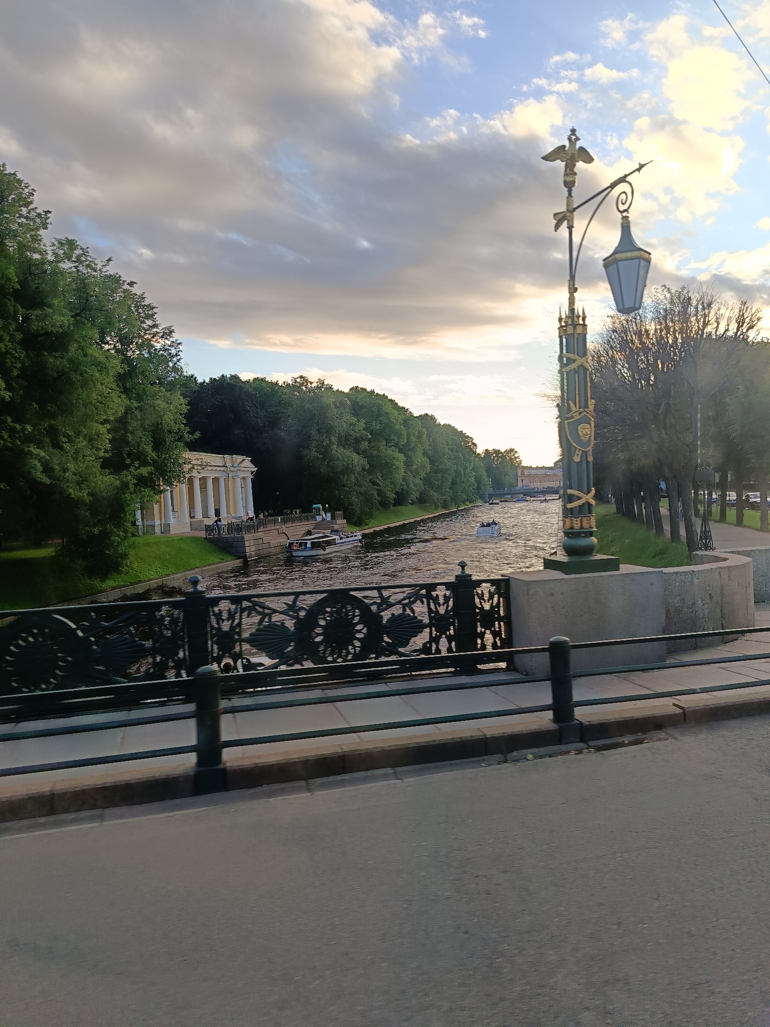 The image depicts a view of the Moyka River in Saint Petersburg, Russia, during what appears to be late afternoon or early evening. In the foreground, a pedestrian bridge railing made of dark metal with intricate scrollwork is visible, suggesting a location with historical architecture. Beyond the bridge, the Moyka River flows, with several small boats navigating its waters. On the left bank of the river, a neoclassical pavilion with white columns and a light-colored roof is situated, surrounded by lush green trees. The right bank features a tree-lined promenade and a decorative lamppost with an eagle finial and gold accents, indicative of Saint Petersburg's elaborate street furniture. The sky above is partly cloudy, with sunlight breaking through, creating a dramatic lighting effect on the scene. The overall impression is one of a serene and picturesque urban landscape within a historically rich city.