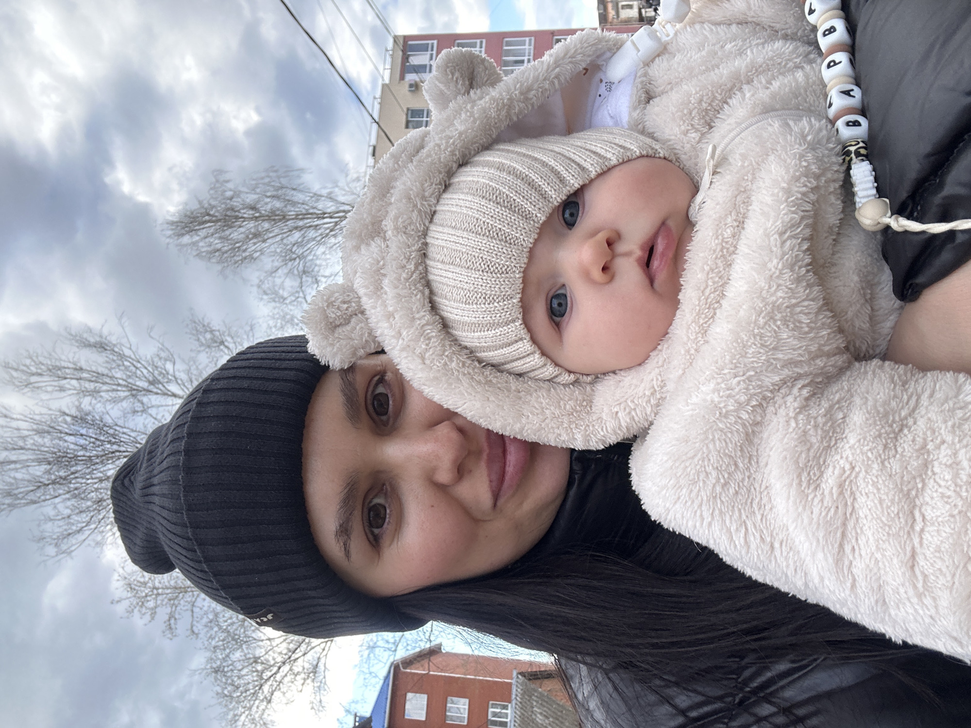 A close-up, rotated outdoor shot features a woman holding a baby in what appears to be Chaikovsky, Russia. The woman, positioned towards the lower left, has dark hair, dark eyes, and a subtle smile. She wears a black ribbed beanie and a dark, possibly leather or puffer, jacket.
The baby, held close by the woman and positioned towards the upper right, is bundled in a warm cream-colored fuzzy snowsuit with bear-like ears on the hood, and a matching knitted beanie. The baby has striking blue eyes and looks directly at the camera with a curious expression.
In the background, a cloudy sky dominates the upper half of the image. Bare tree branches are visible against the sky, suggesting late autumn or winter. Several multi-story buildings are seen in the distance; one prominently features reddish-brown facade, while another is lighter in color. Power lines or cables are visible stretching across the sky.
A notable detail is a beaded accessory, likely a pacifier clip or teether, hanging from the baby's clothing. It features square beads with individual letters, which appear to spell "L O P A P B L". The overall impression is a tender moment between a caregiver and child on a cool, overcast day.