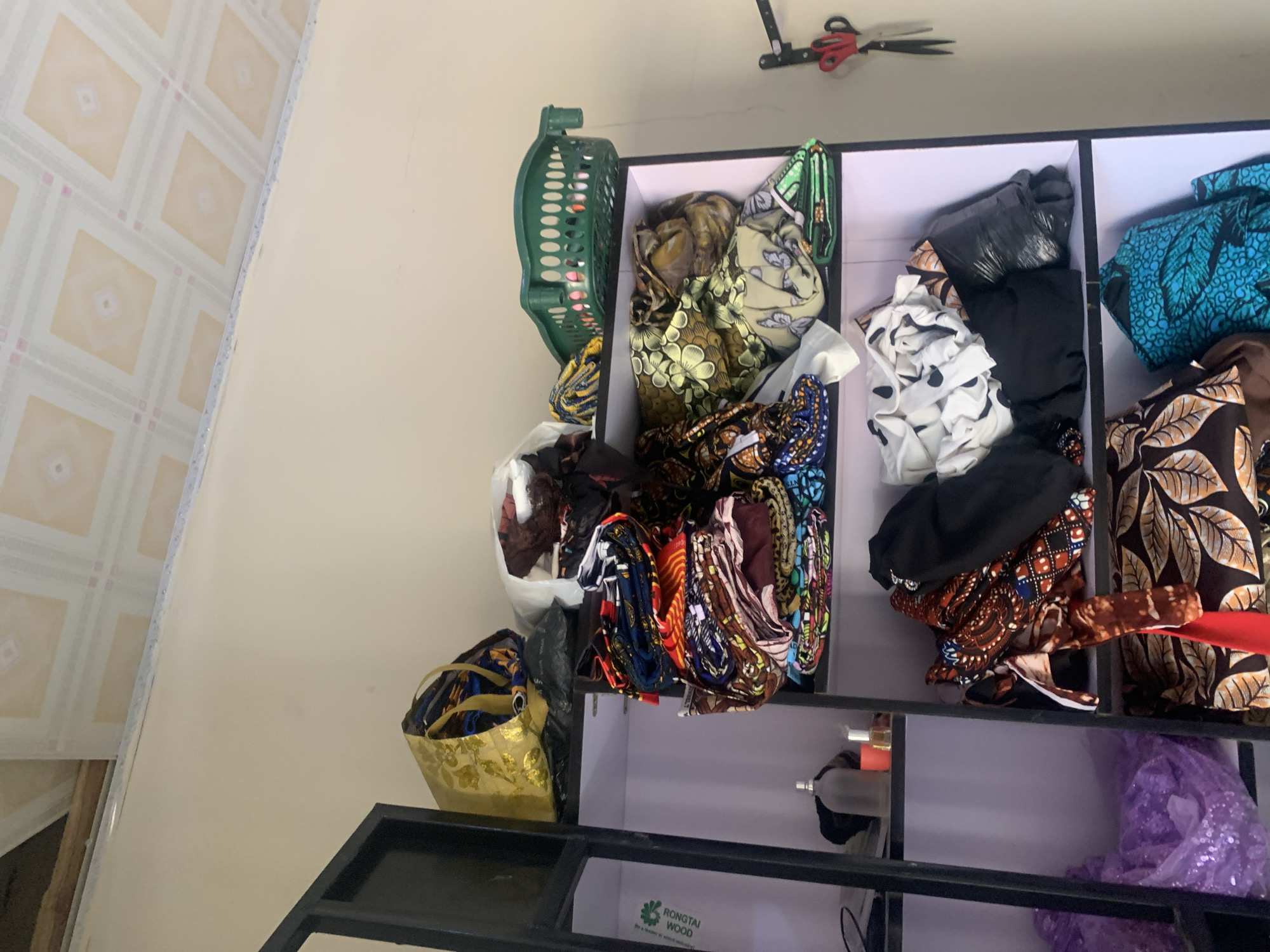 The image depicts a shelving unit filled with fabrics and other items, likely in a storage area or shop in Jauro Yinu, Nigeria. The top shelf holds a gold shopping bag with fabric peeking out, a black plastic bag, and a white bag containing more fabric. To the right, a green plastic basket sits on the shelf, containing fabric. The middle shelves are stacked with colorful, folded fabrics, some with intricate patterns. The lower shelves show more folded fabrics, including some with leaf patterns and a bright blue fabric with a pattern. On the far left, a clear bottle and a small orange container are visible on a shelf. The overall environment appears to be indoors, with a tiled ceiling visible at the top of the image. A black metal frame, possibly part of a larger structure or furniture, is visible on the left side. A pair of scissors hangs on the wall to the right of the shelving unit.