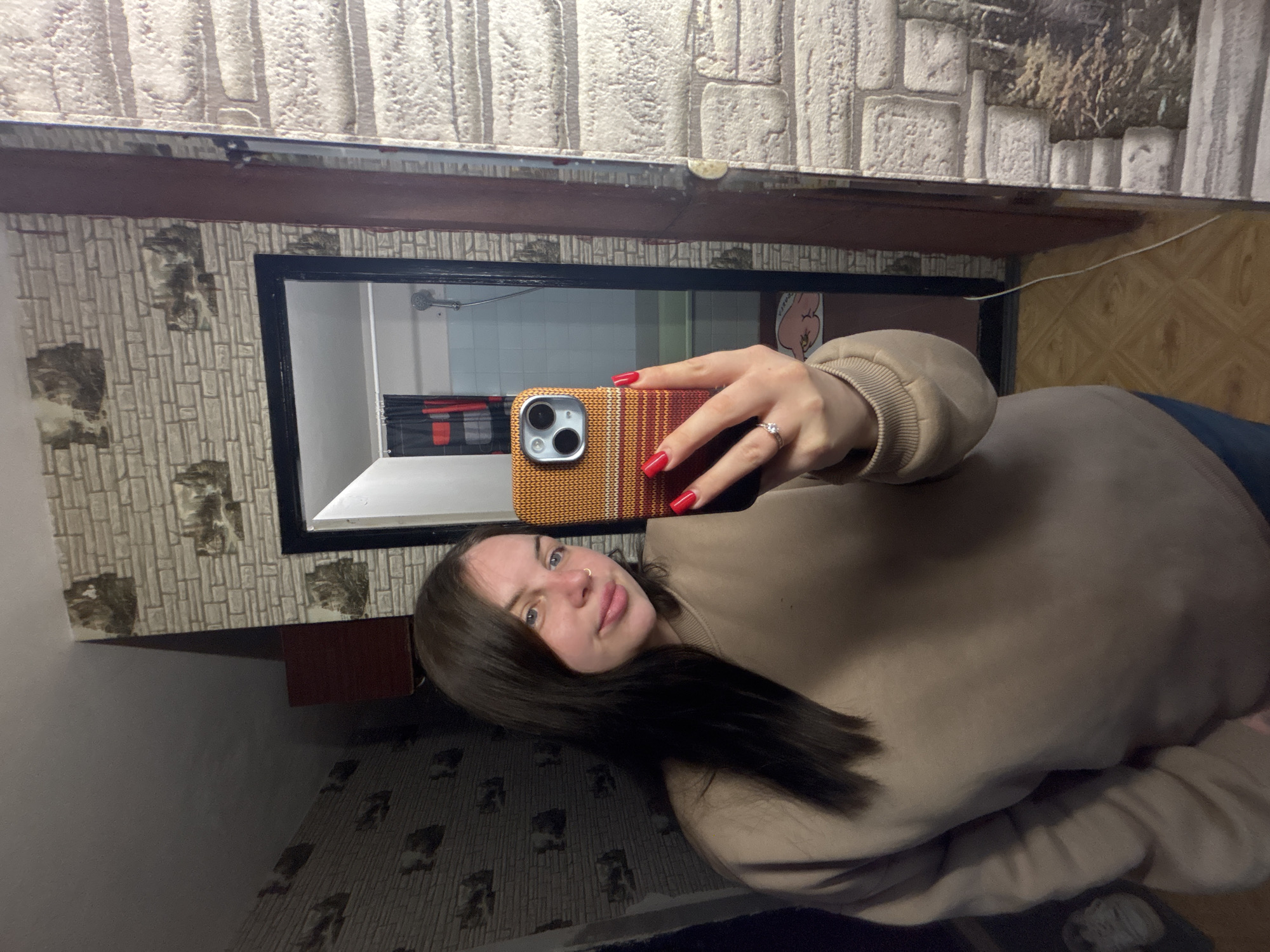 A young woman is depicted taking a mirror selfie, with the image rotated 90 degrees counter-clockwise. She has long, dark hair and light eyes, with a nose piercing and a neutral to slightly pouting expression. She is wearing a light brown or beige sweatshirt and displays red nail polish on her fingers, along with a ring on her left ring finger. Her iPhone is encased in a distinct orange, red, and yellow woven-patterned case.
The background shows an indoor setting, likely a residence in Chornomorskoe, Ukraine. The wall features patterned wallpaper resembling light-colored stone or brickwork, with some areas appearing darker or distressed. A dark wooden beam or shelf is visible above the mirror frame. The mirror reflects a different room, possibly a bathroom, featuring white tiled walls, a visible shower head, and a dark shower curtain or blind. A small, cartoonish pink pig figure is partially visible on a surface within the reflection. The floor, both in the reflection and beneath the woman, is a patterned wooden surface. A white electrical cord hangs in the lower right area of the reflected image. The lighting appears to be artificial, typical of an indoor environment.